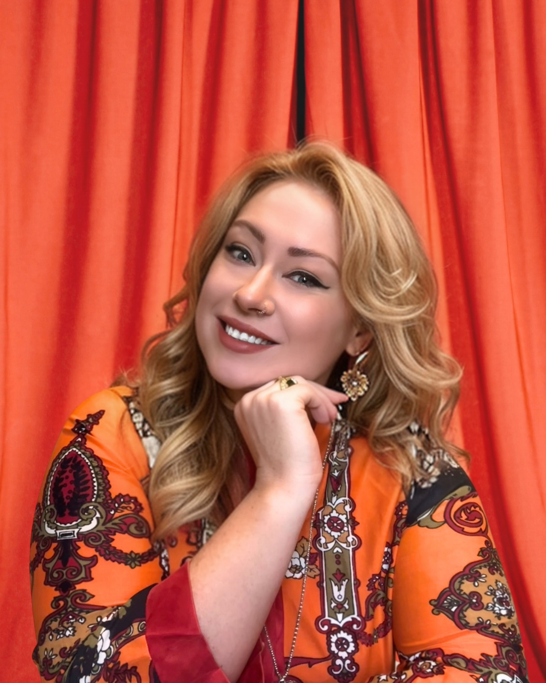 Young woman with curly blonde hair wearing orange patterned blouse, smiling, posing in front of red curtains.