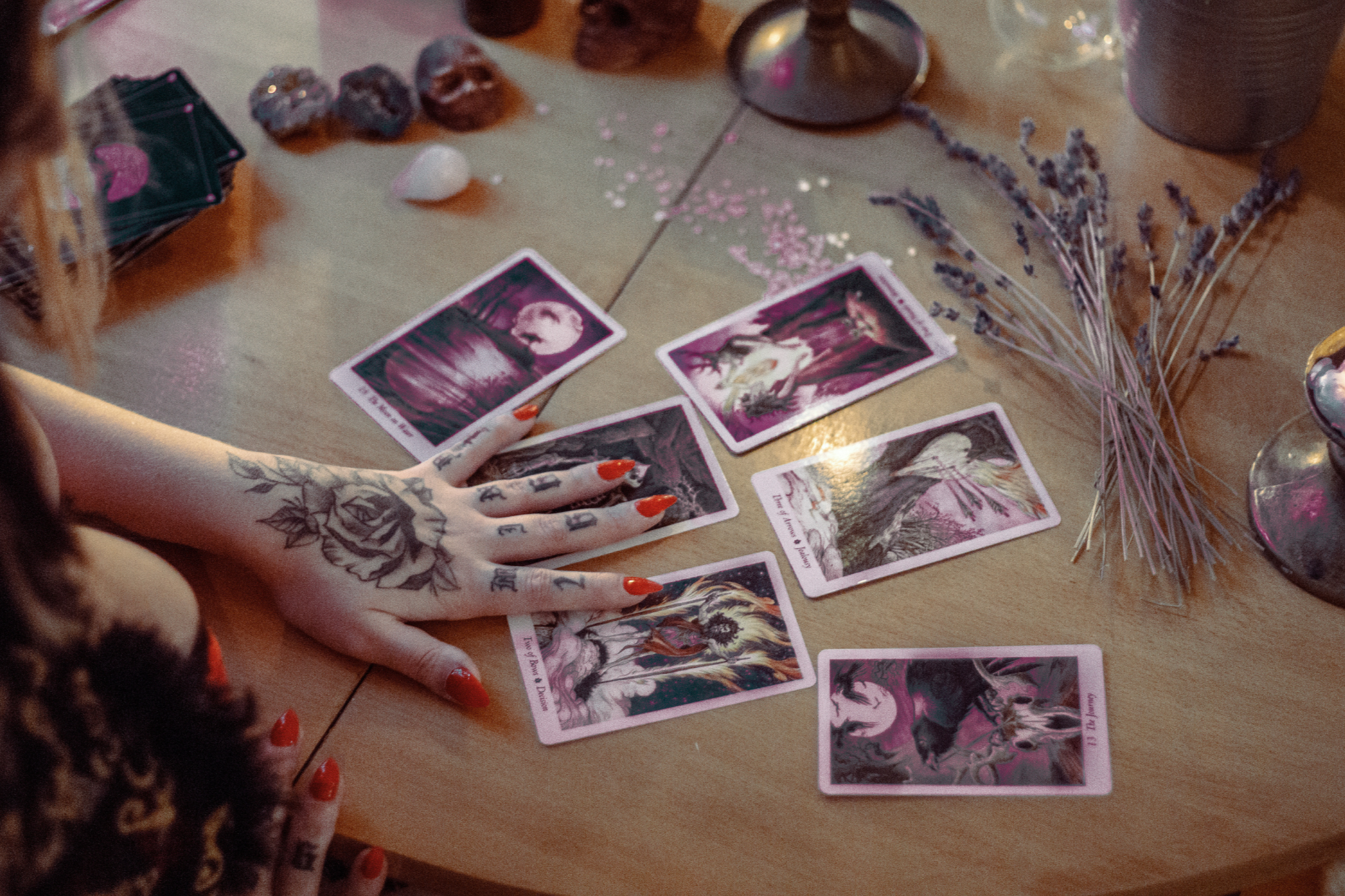 A person with decorated hands, tarot cards spread on a wooden table, including lavender sprigs, crystals, candles, and decorative items.
