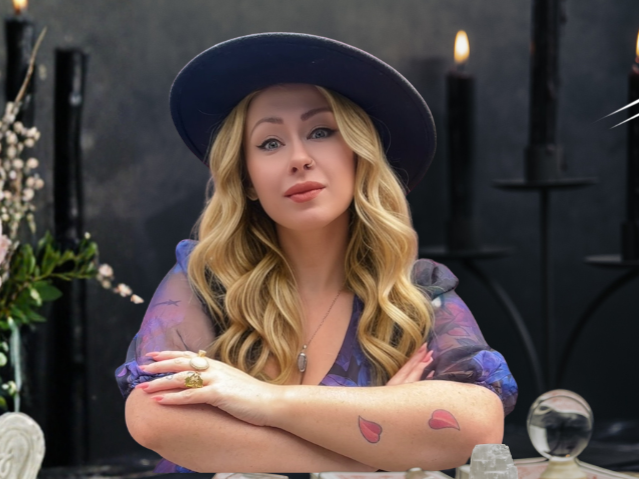 A woman with long blonde hair wearing a purple hat and purple dress, sitting at a table with candles and a glass orb, with a dark background and candlelit ambiance.