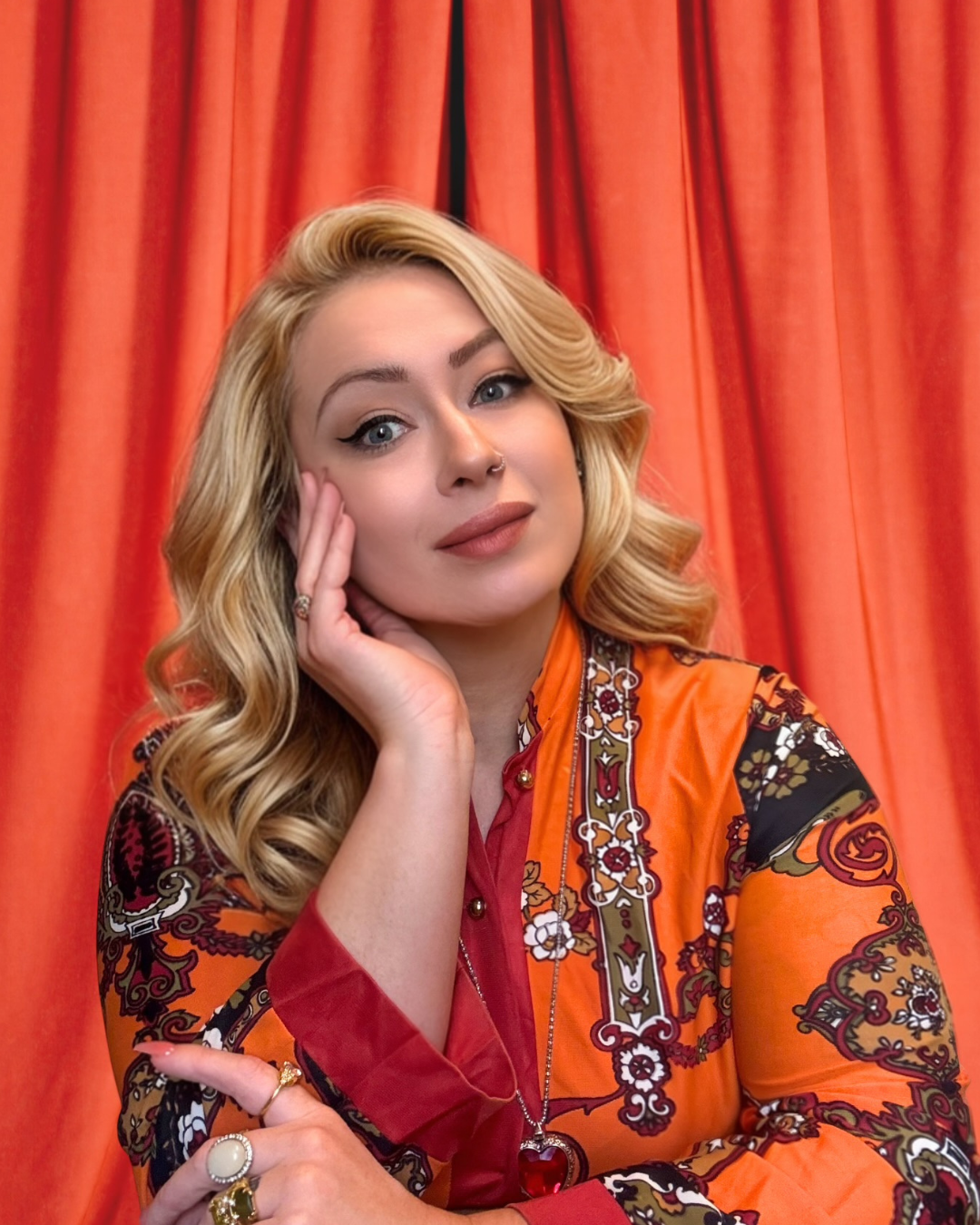 Portrait of a woman with wavy blonde hair, makeup, and jewelry, standing in front of red curtains. She looks like she is magical