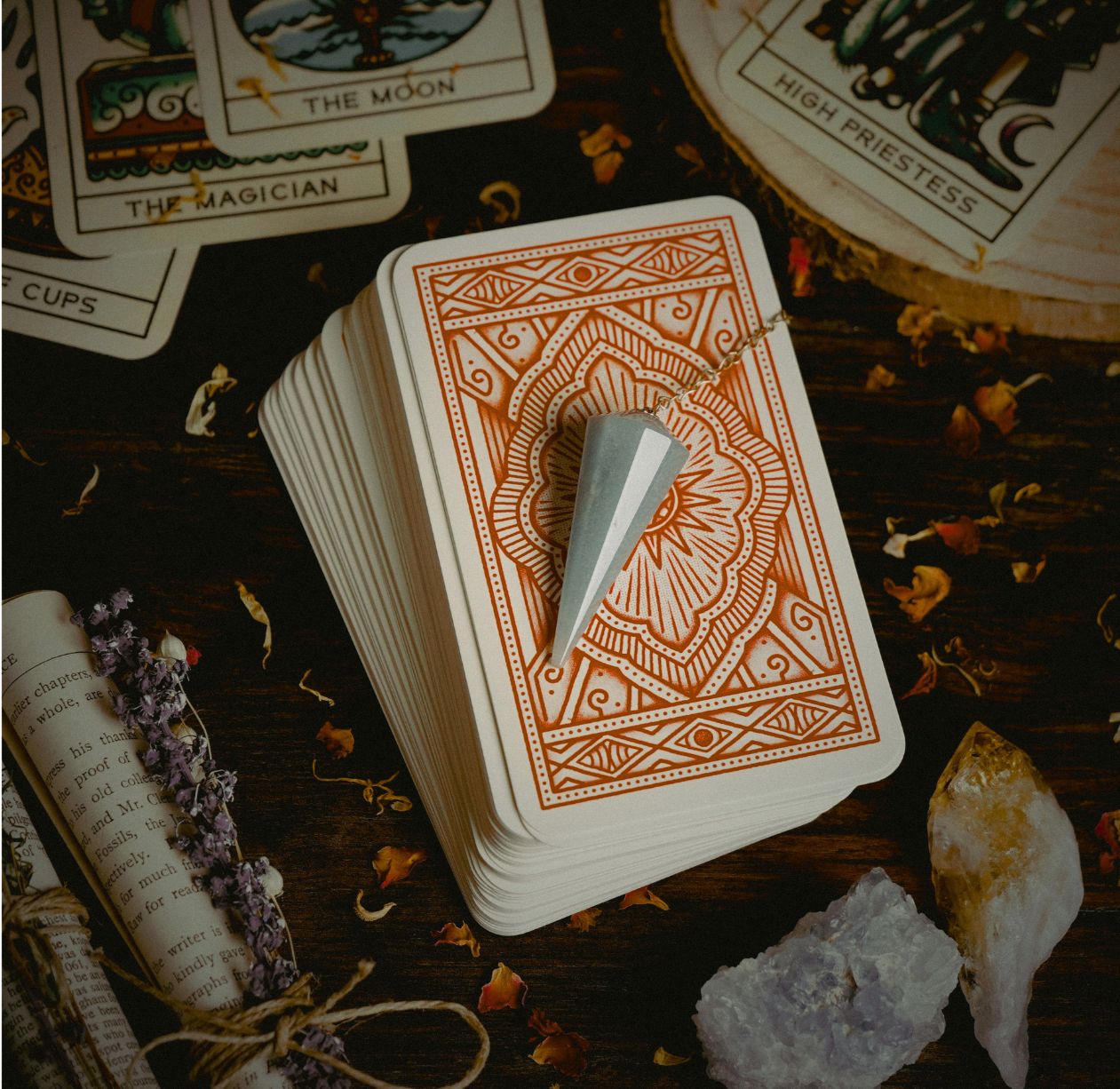 A deck of tarot cards with an orange and white intricate back design is laid out on a dark wooden surface. A large, pointed crystal wand rests on the deck. Around the deck are scattered flower petals, a sprig of dried purple flowers, a rolled-up book, a yellow quartz crystal, and other smaller stones.