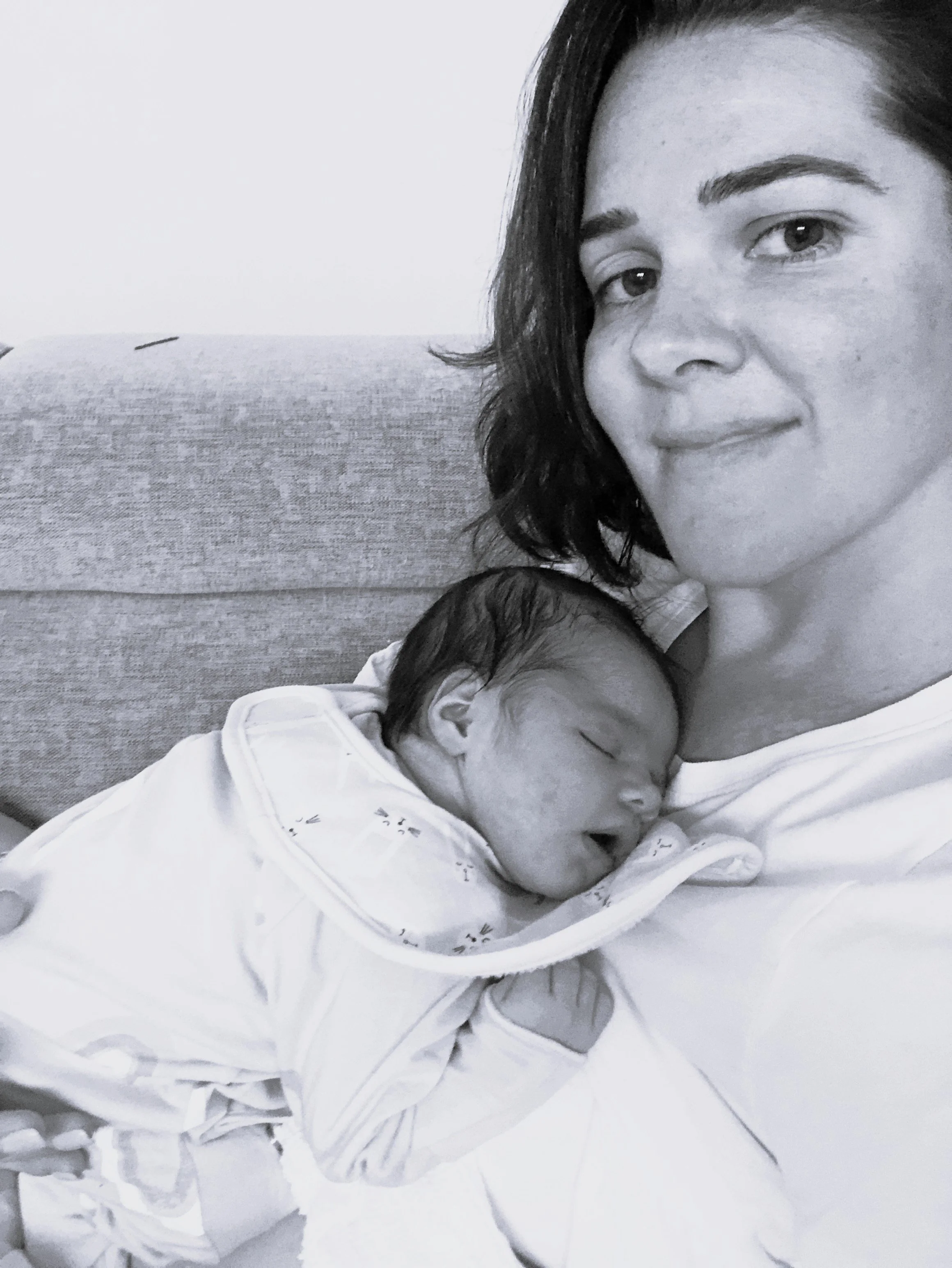 A woman with dark hair and light skin is resting on a couch, with her eyes open and a gentle smile. A newborn baby swaddled in white is sleeping on her chest.