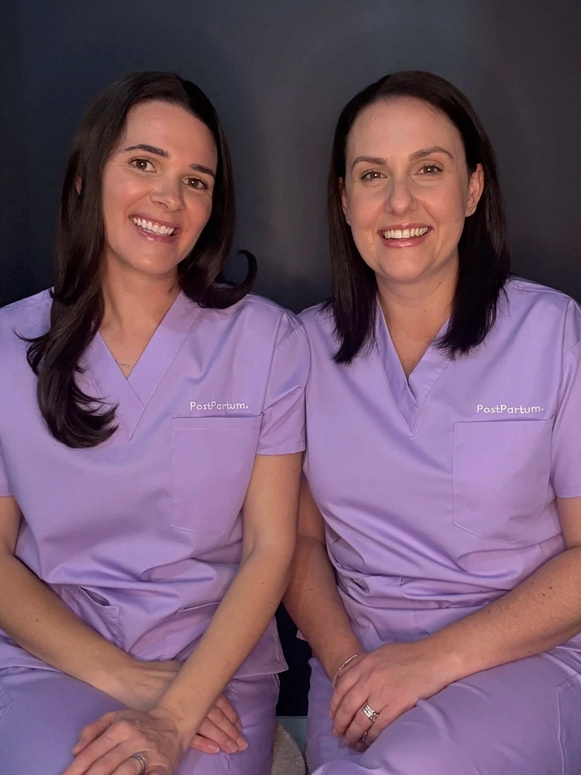 Two PostPartum midwives in purple scrubs, seated together and smiling at the camera.