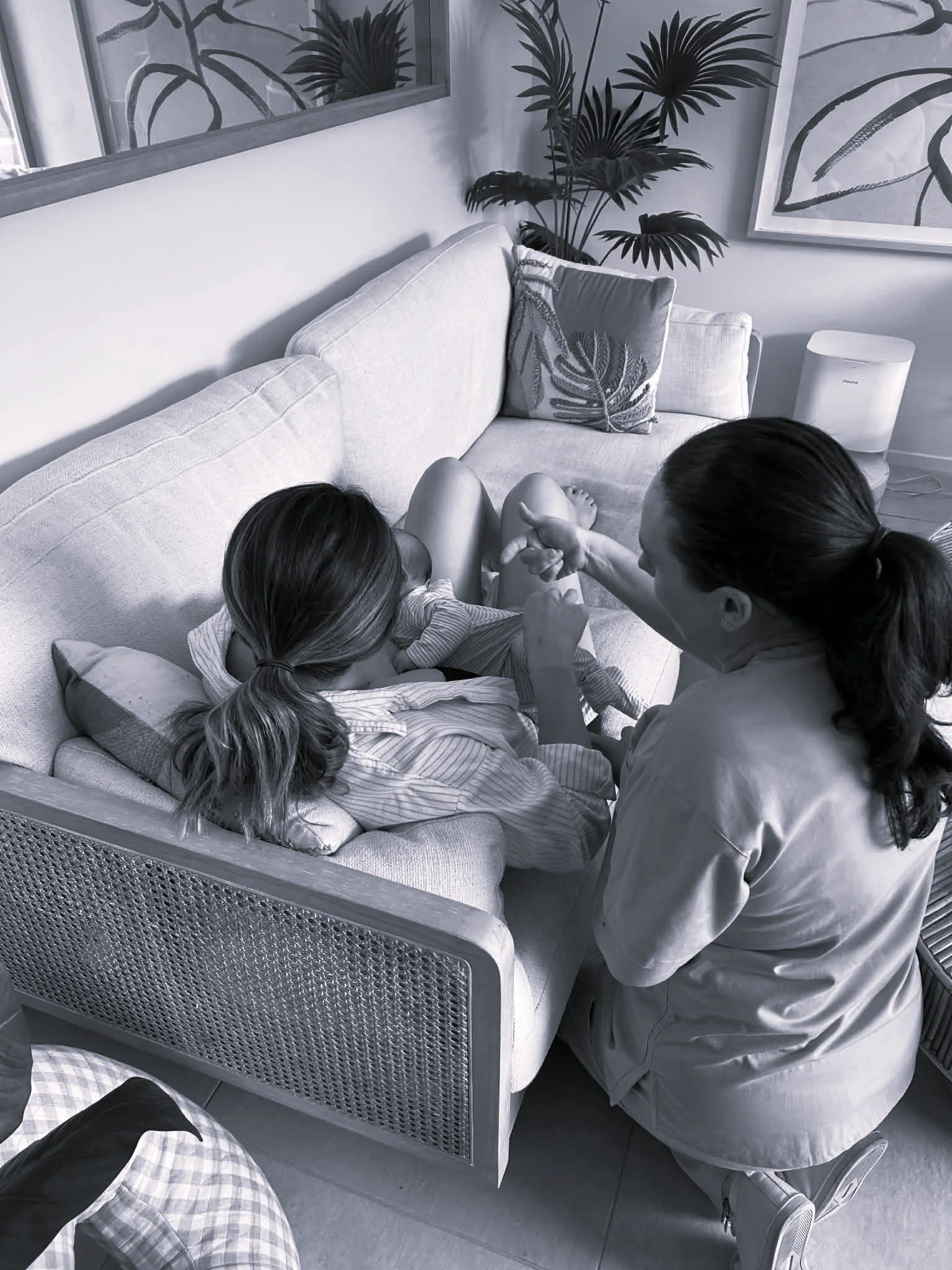A woman with glasses sitting on a sofa with a child, and another woman with glasses and braided hair crouching nearby. The room has tropical plant artwork and a large potted plant.