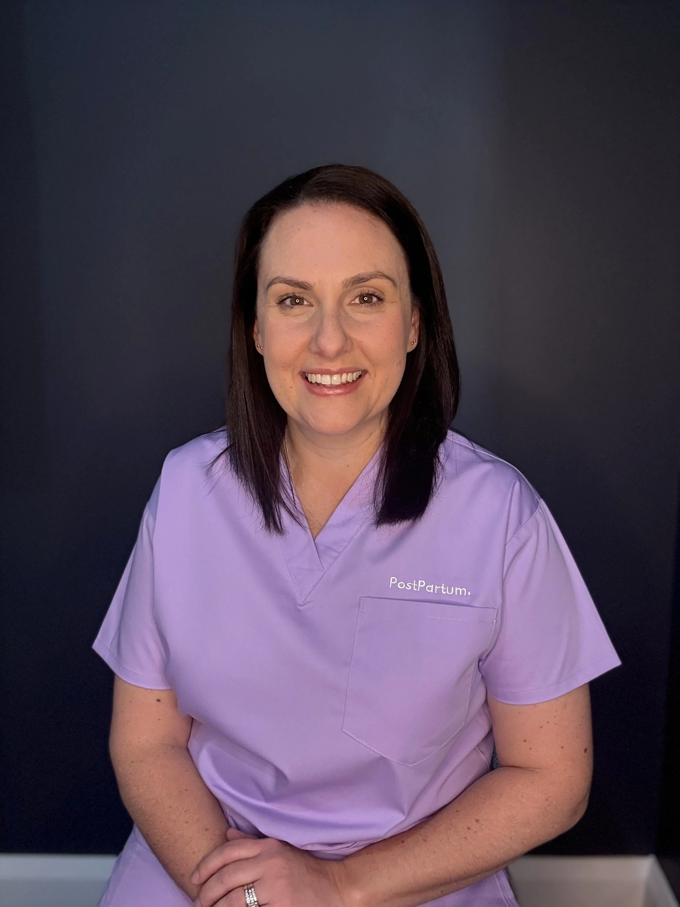 A woman with dark brown hair, smiling, wearing a light purple medical scrub with 'PostPartum' embroidered on it, standing against a dark background.
