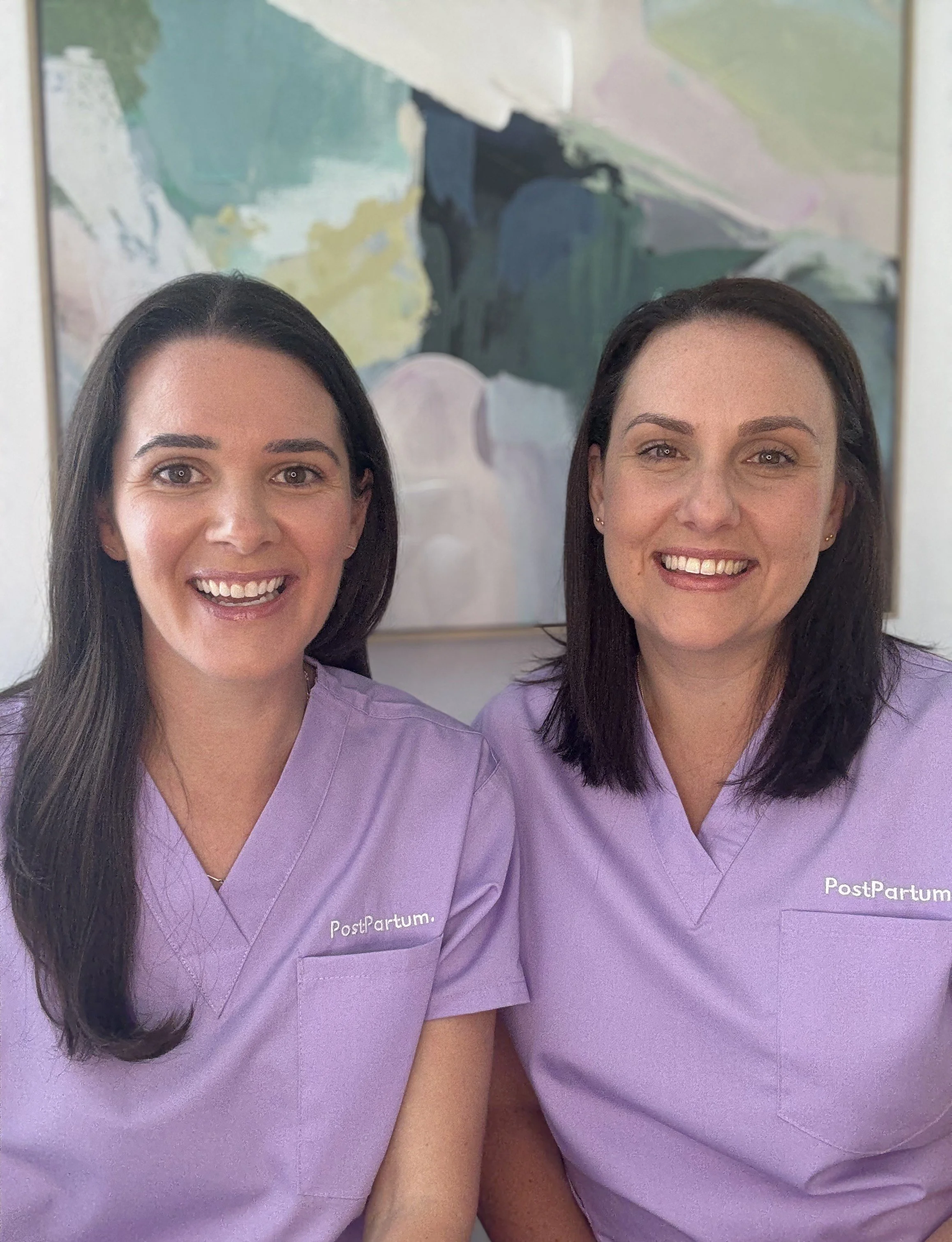 Two PostPartum midwives in purple scrubs, smiling together in front of a colourful artwork.