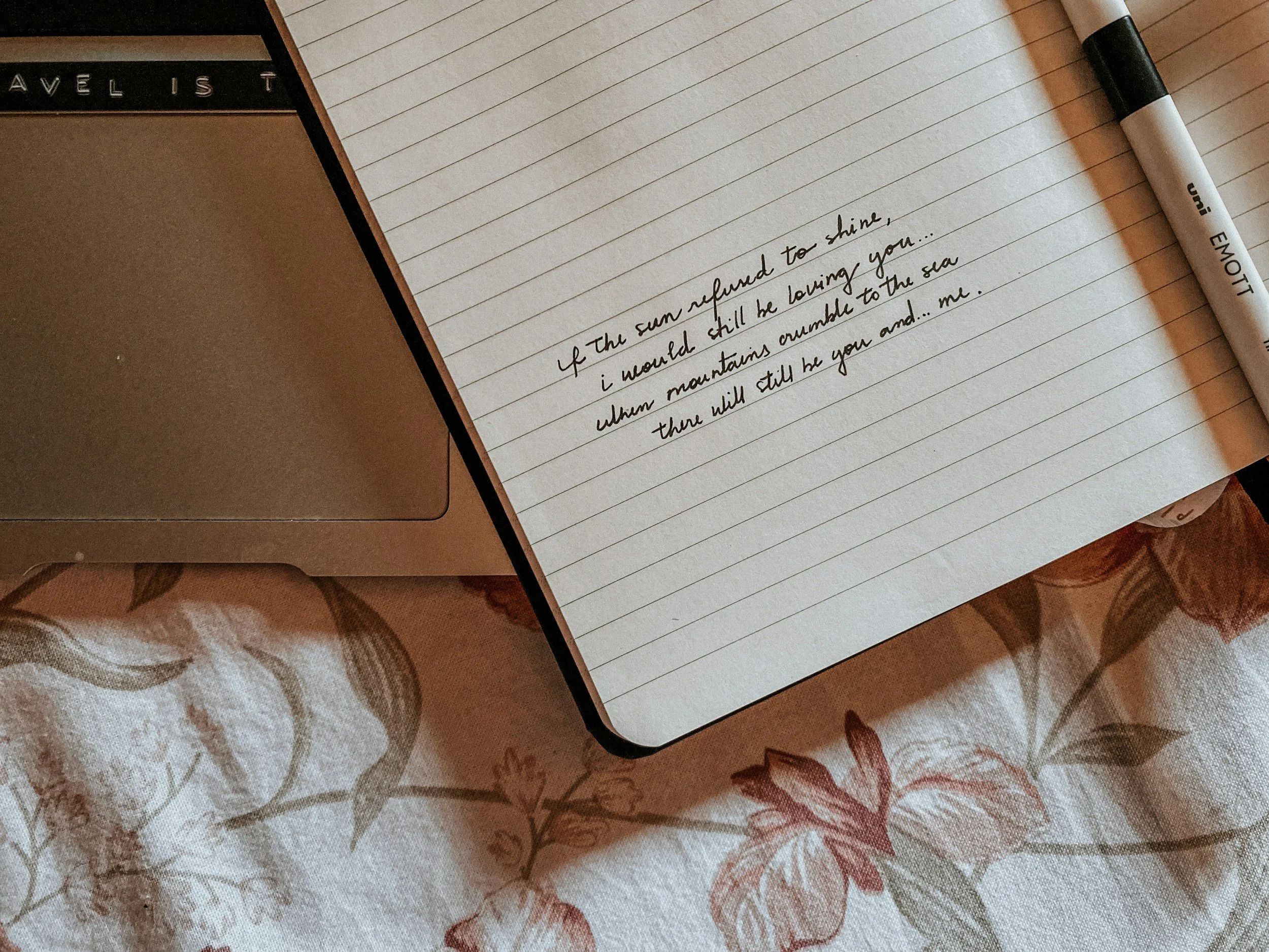 Open notebook with handwritten poem, a pen labeled 'Emoti,' and a laptop on floral tablecloth.
