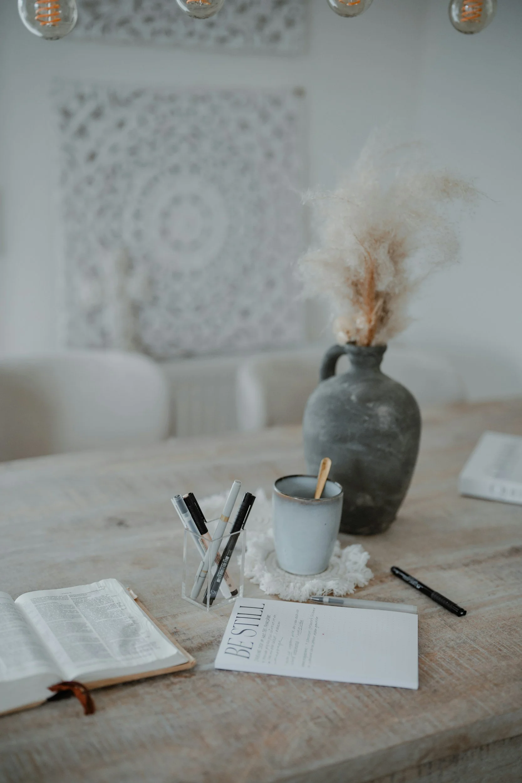 A wooden table with an open Bible, a cup with pens, a drink with a spoon, a large ceramic vase with dried pampas grass, and a paper titled 'BE STILL' in a cozy, well-lit room.