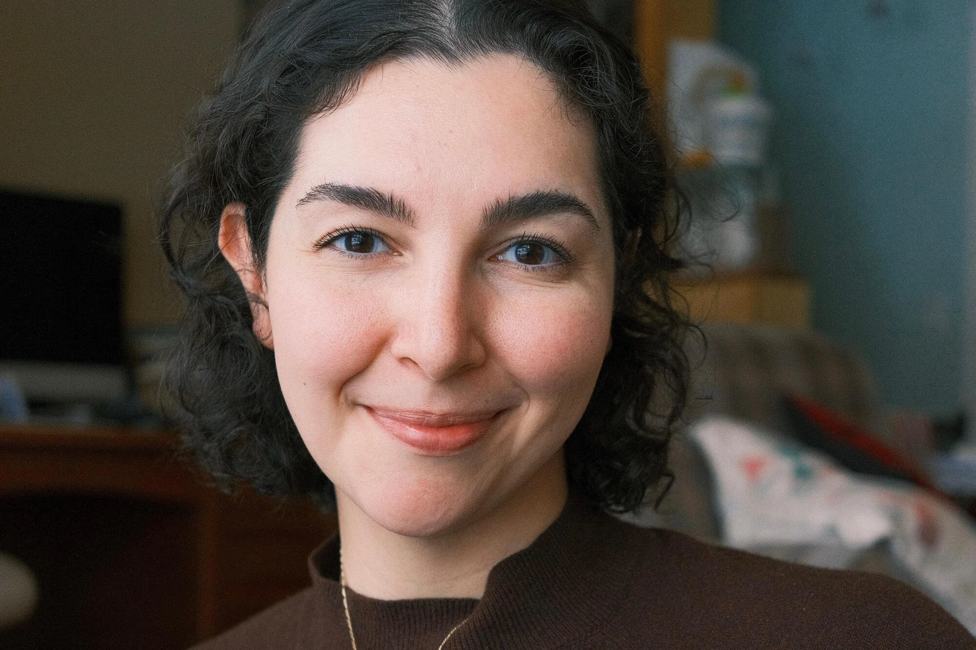 Close-up of a Alia smiling. She has dark, curly hair and light skin in an indoor setting.