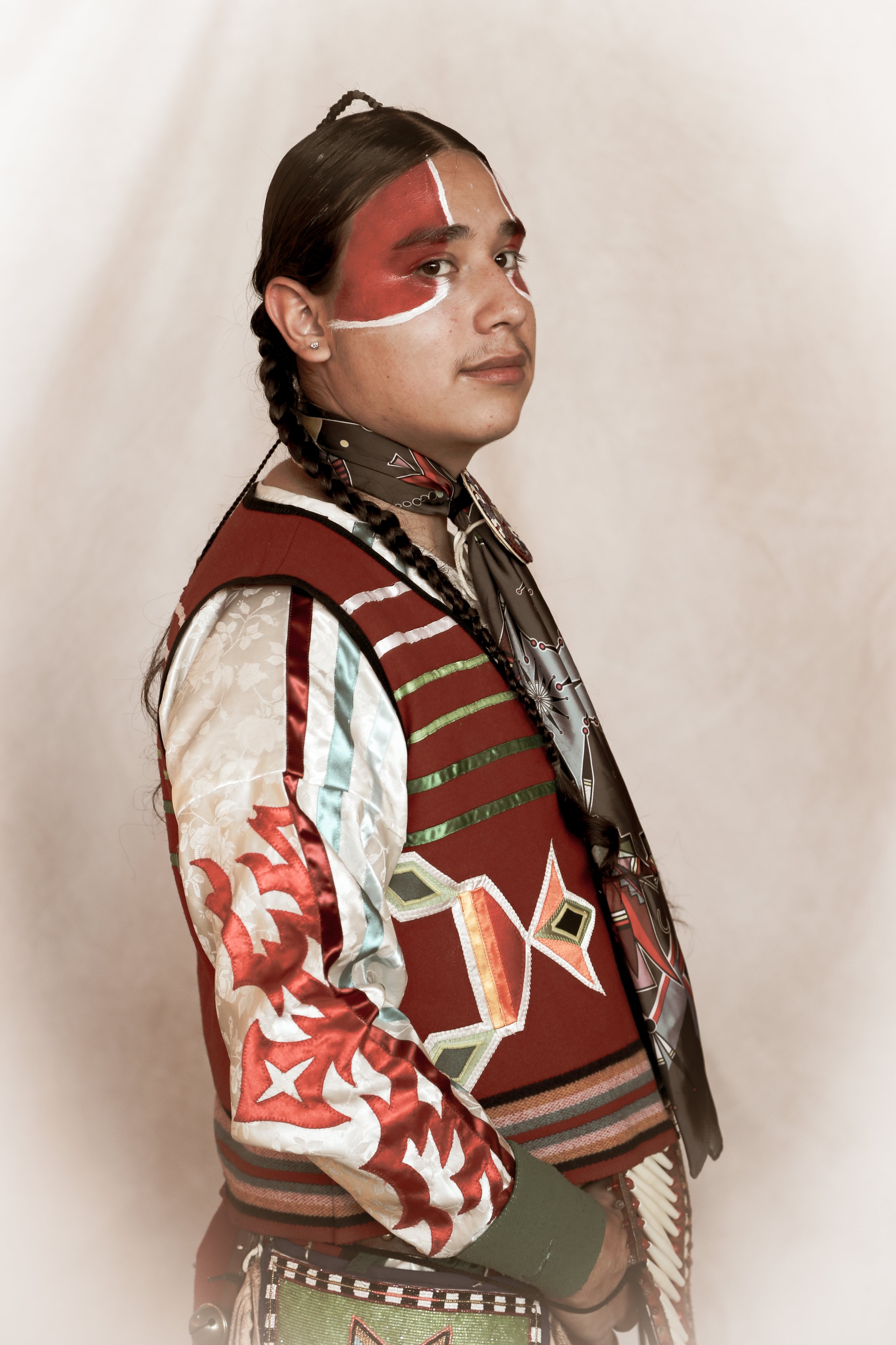 A person with braided hair wearing traditional, colorful attire and face paint, looking to the side. The attire includes intricate patterns and designs.