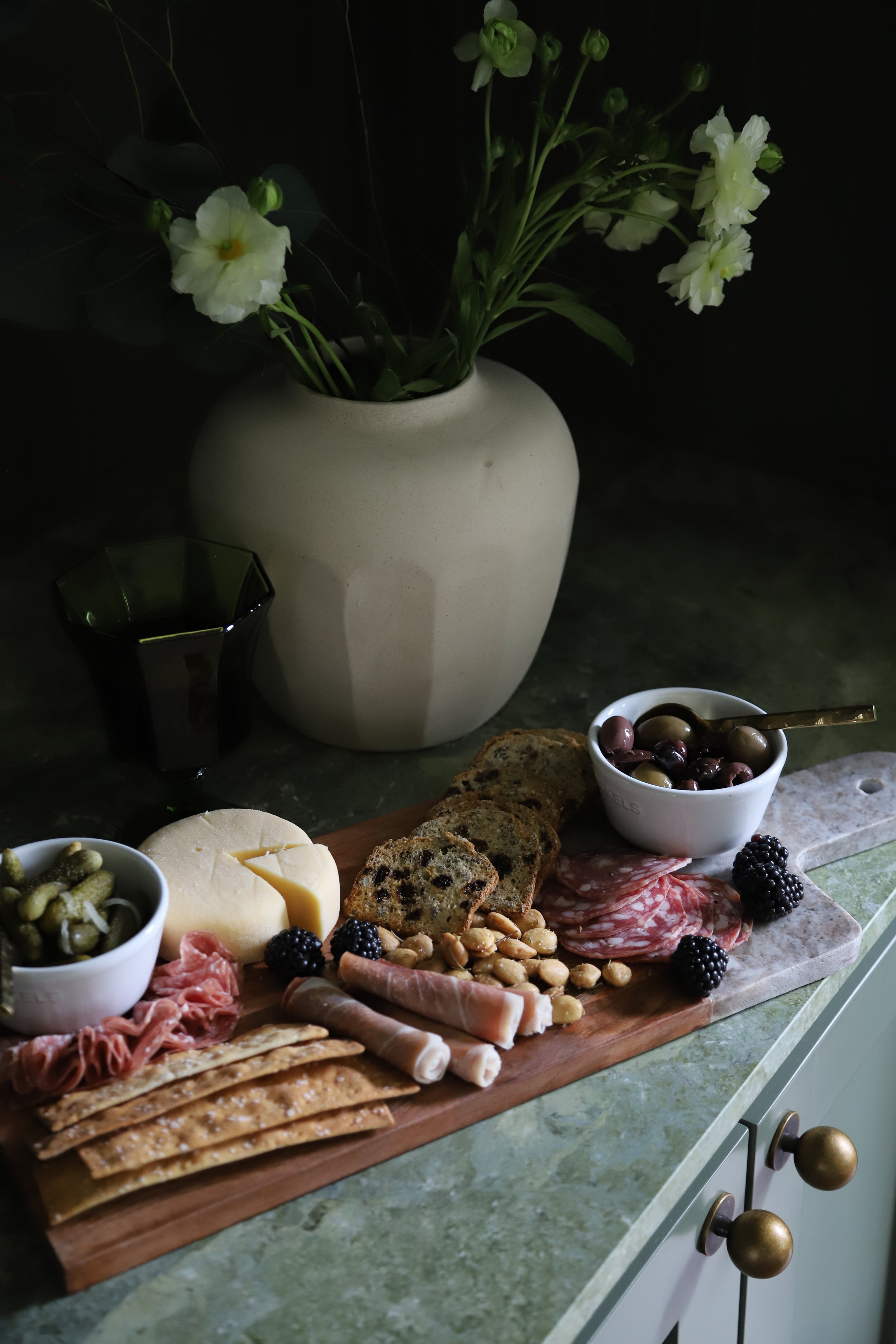 Charcuterie board with cheese, cured meats, crackers, olives, blackberries, and nuts on a marble countertop, with a vase of white flowers and a dark glass nearby.