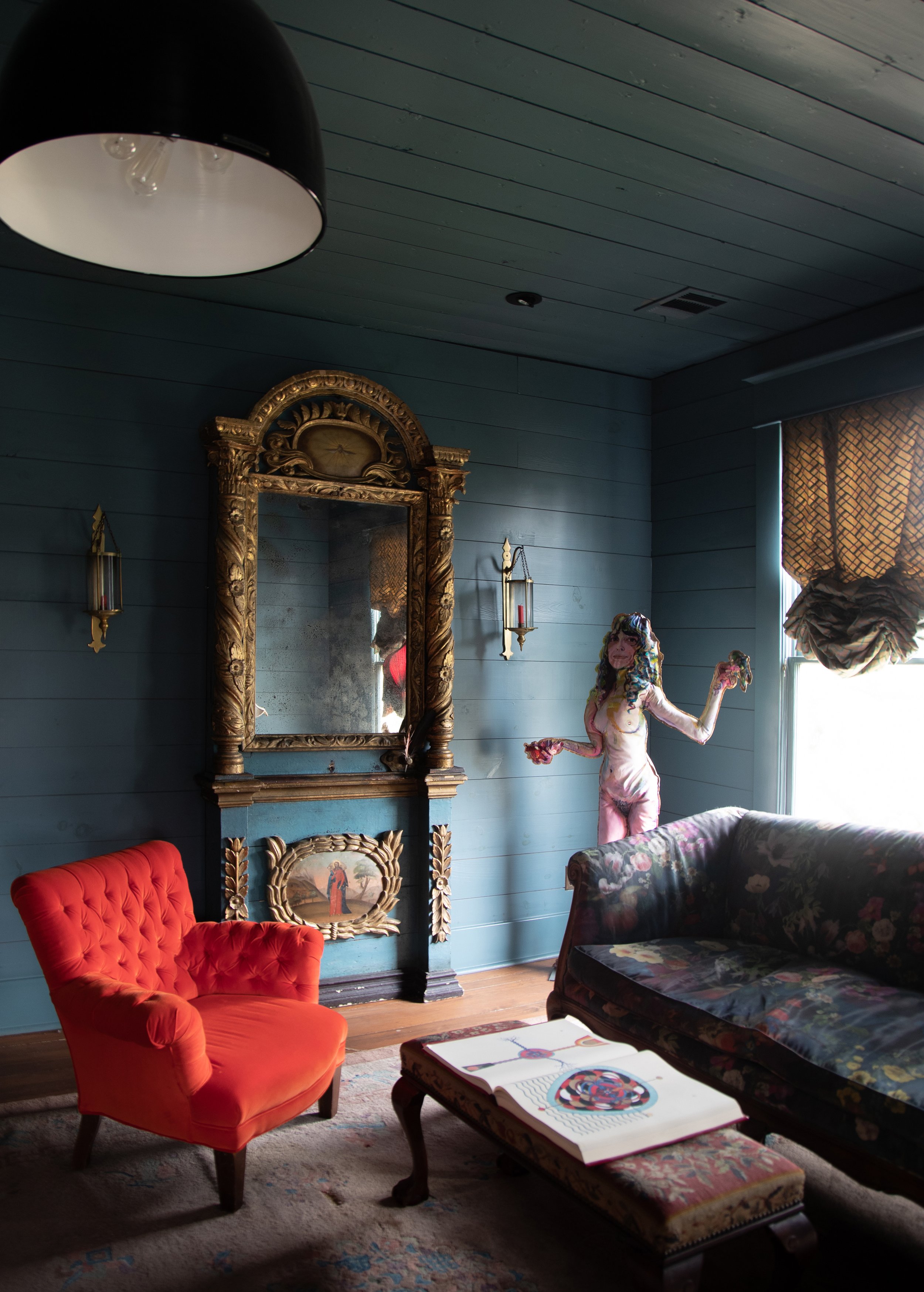 A cozy living room with blue wooden walls, featuring a red tufted armchair, a floral-patterned couch, and a vintage mirror with ornate gold detailing. A whimsical, colorful statue stands near the window, and a patterned ottoman with a book is in the