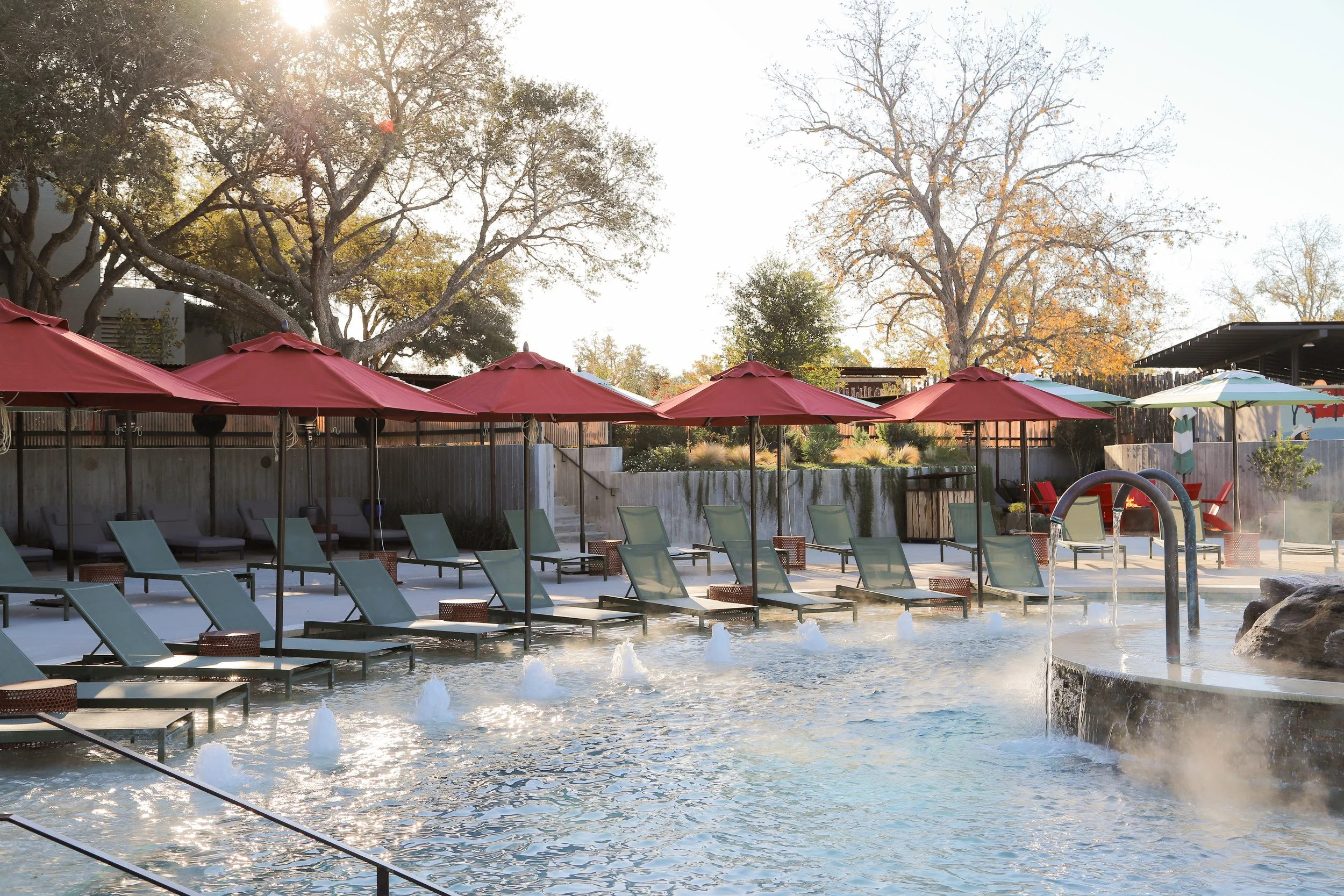Swimming pool with multiple lounge chairs and red umbrellas, surrounded by trees and a wooden fence, during daytime.