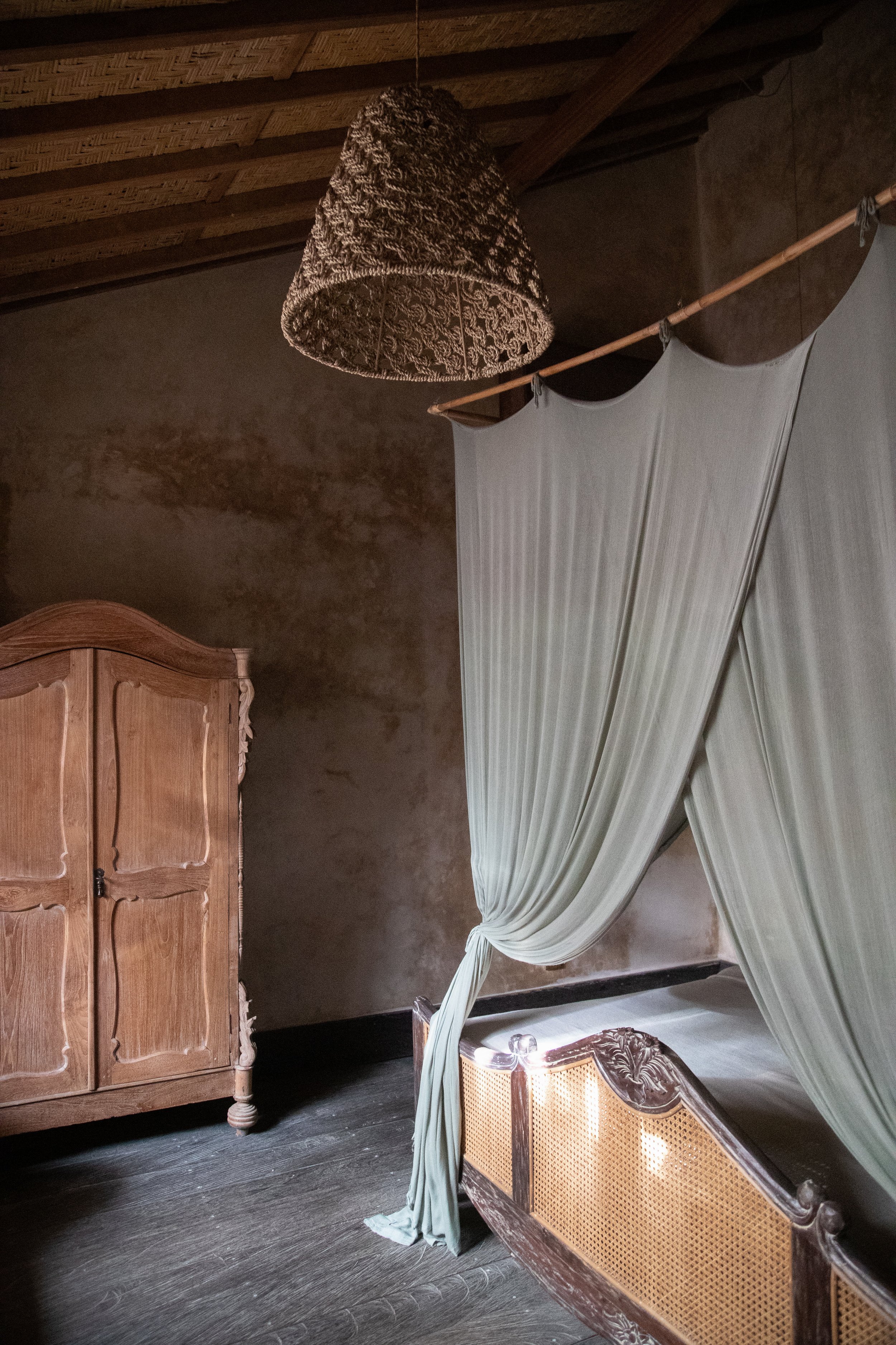 Rustic bedroom with a wooden wardrobe, canopy bed with draped fabric, and a woven lampshade.