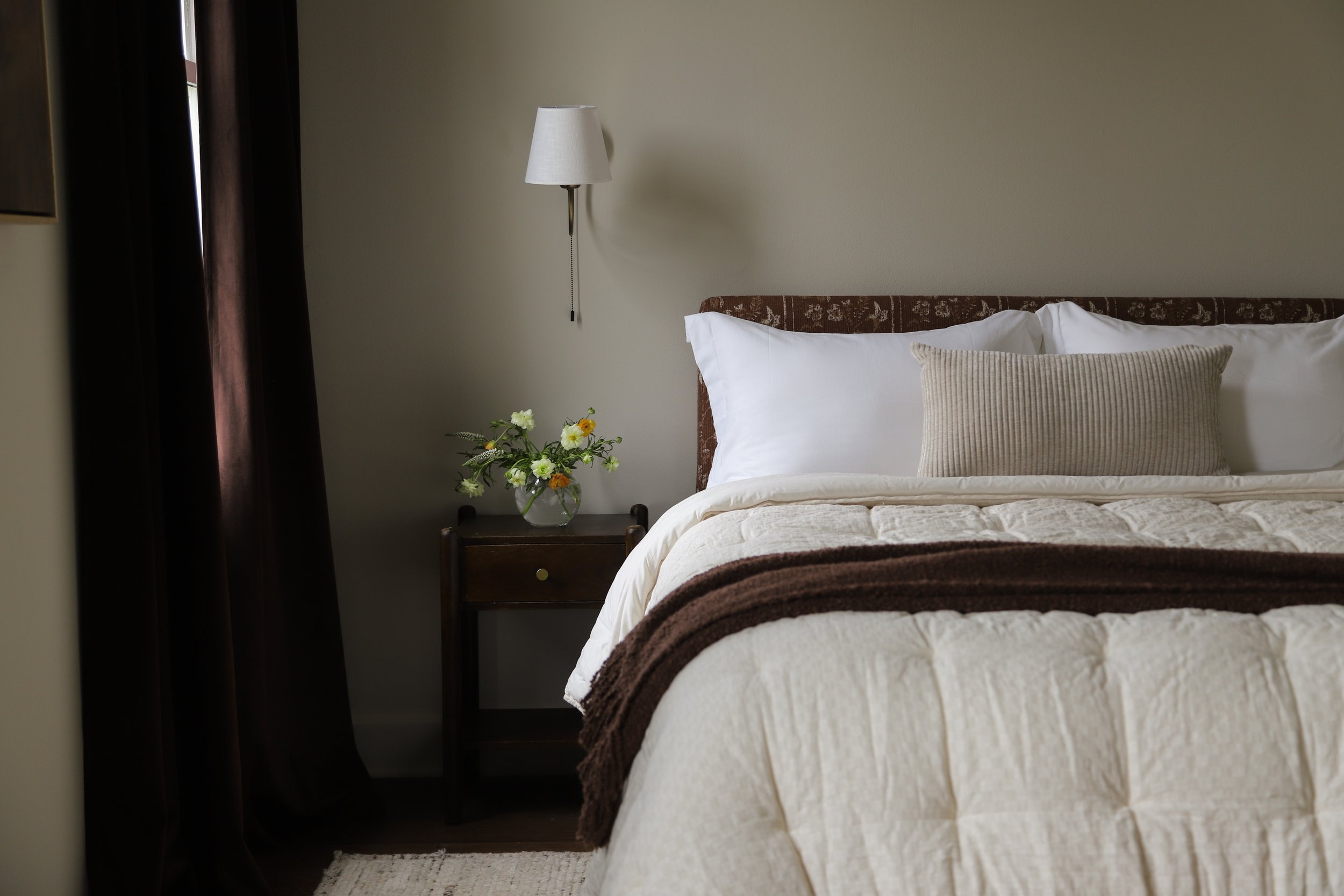A neatly made bed with white linens, a beige pillow, and a brown blanket, next to a wooden nightstand with a glass vase of flowers on it, and a wall-mounted lamp above.