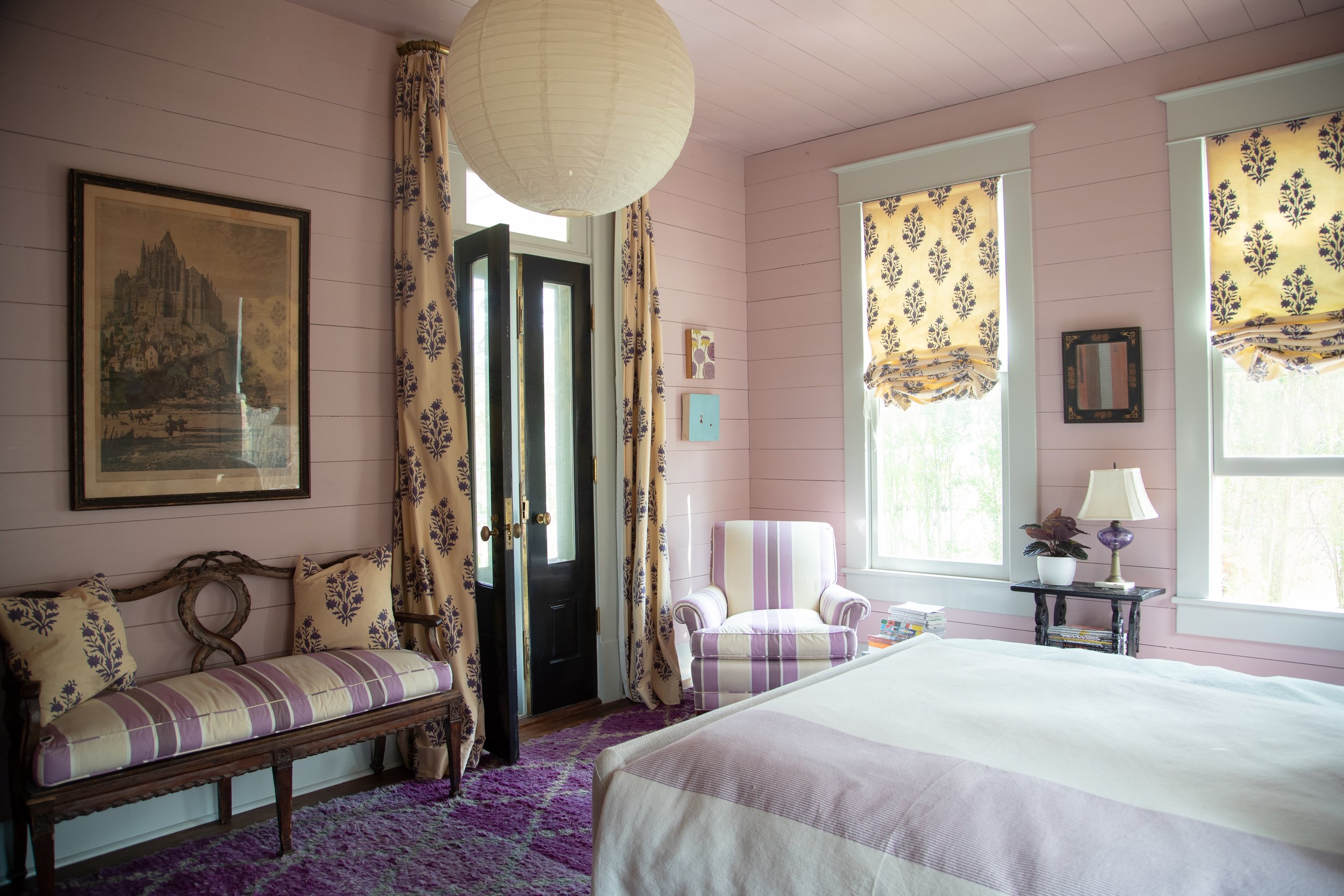 Elegant bedroom with light pink walls and a large hanging paper lantern. The room features a vintage bench with purple and cream striped cushions, matching armchair, and decorative pillows. A framed artwork hangs on the wall. Floral curtains and Roma