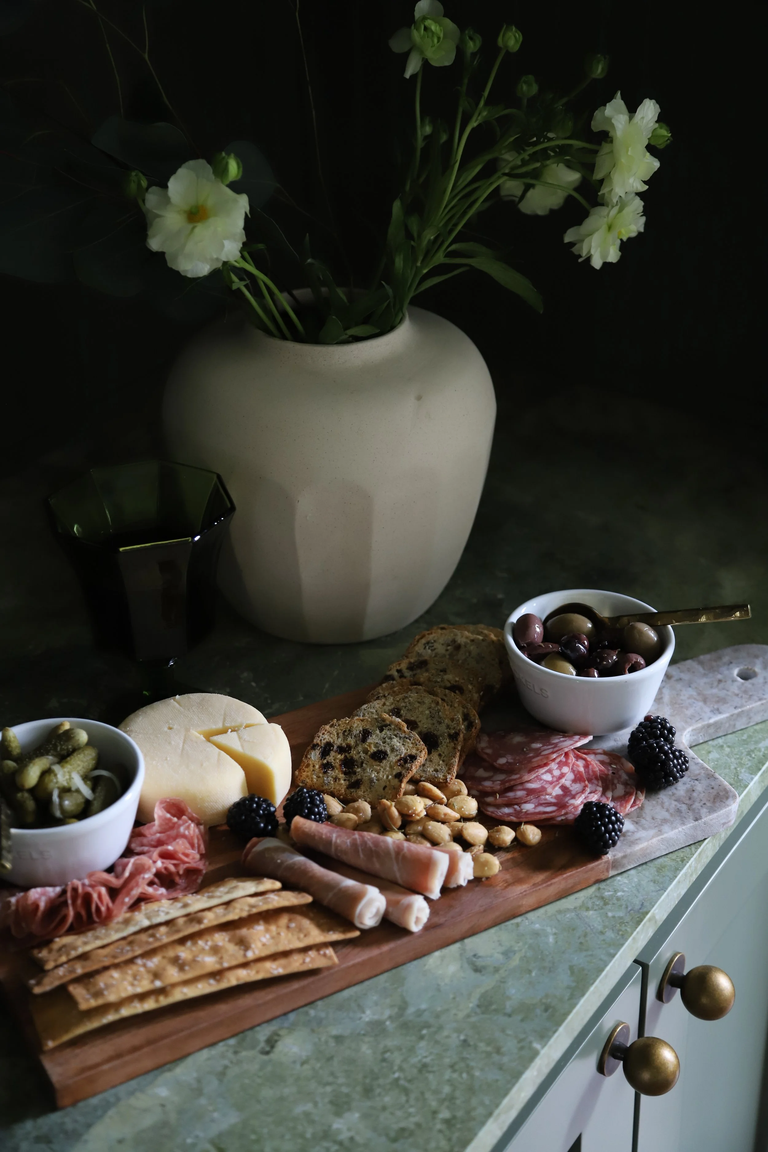A wooden serving board with cheese, cured meats, crackers, olives, blackberries, and nuts, placed on a green marble countertop. In the background, a white vase with white and green flowers and a black glass are visible.