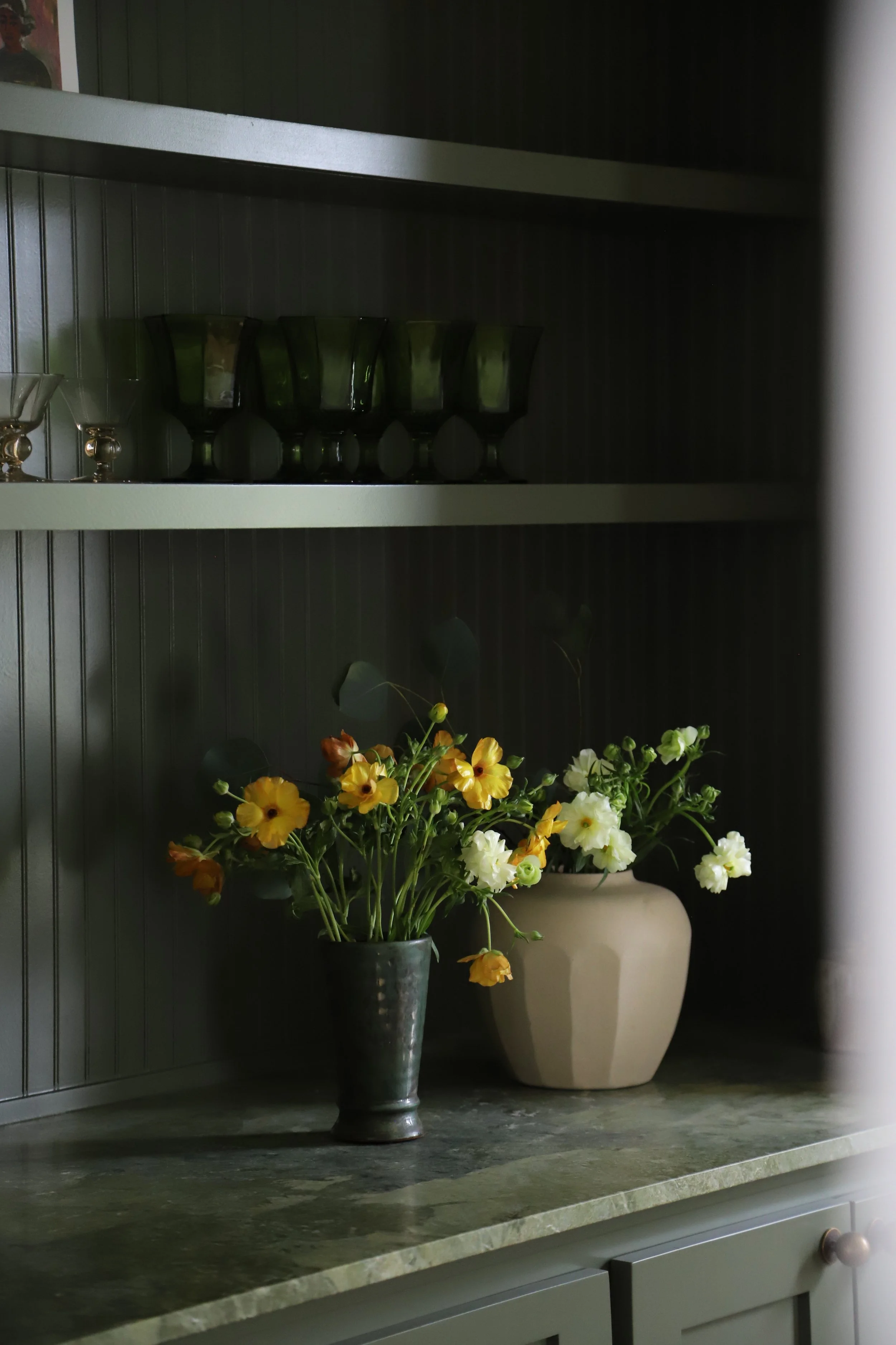 A dark green cabinet with a green marble countertop, two potted flowers—one with yellow and orange flowers in a metallic pot, and the other with white flowers in a beige pot—on the surface, and glassware on the shelves above.