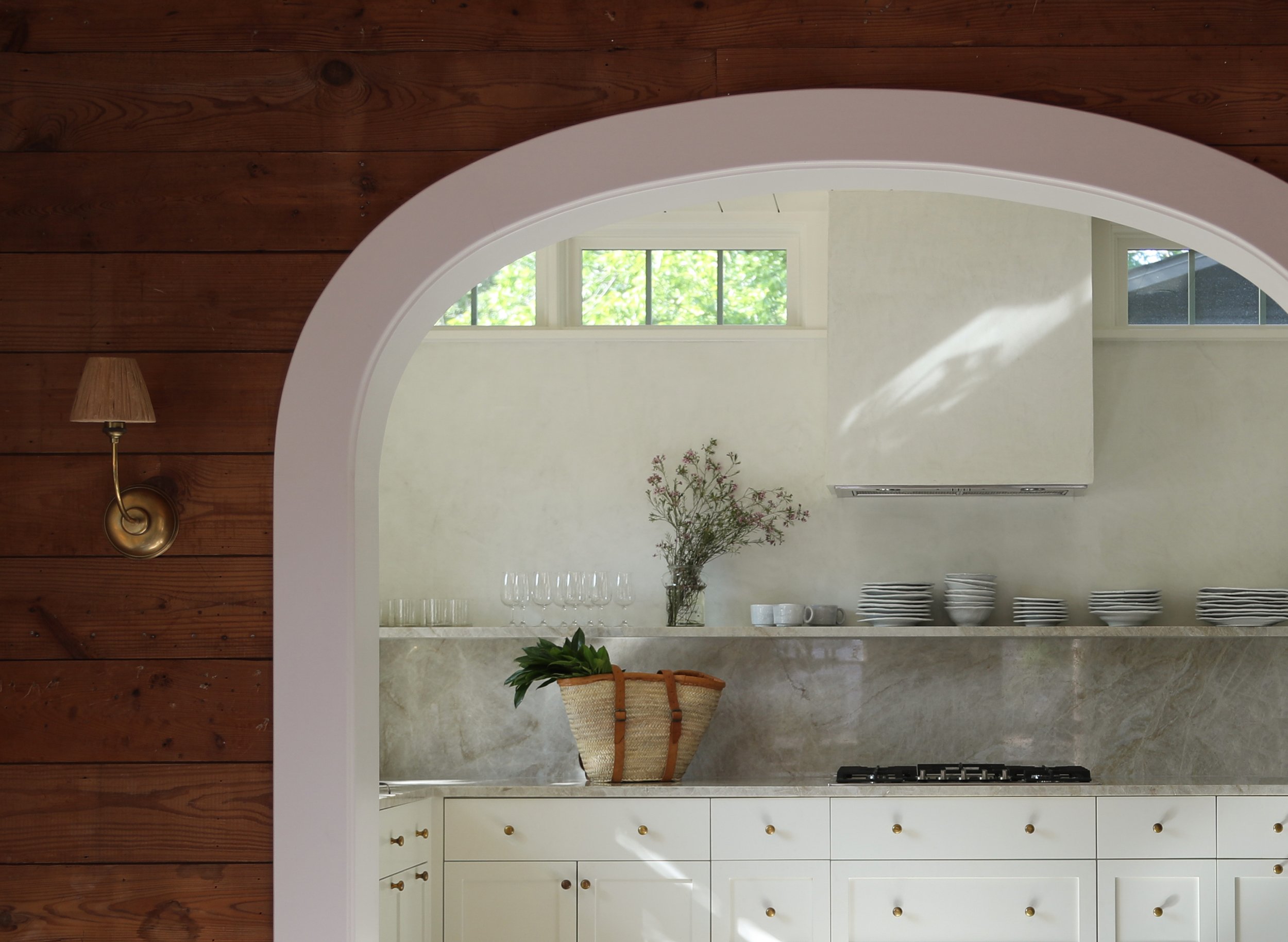 View of a modern kitchen through an arched doorway, featuring white cabinets, a marble countertop, a decorative vase with flowers, and stacked dishes on a shelf.