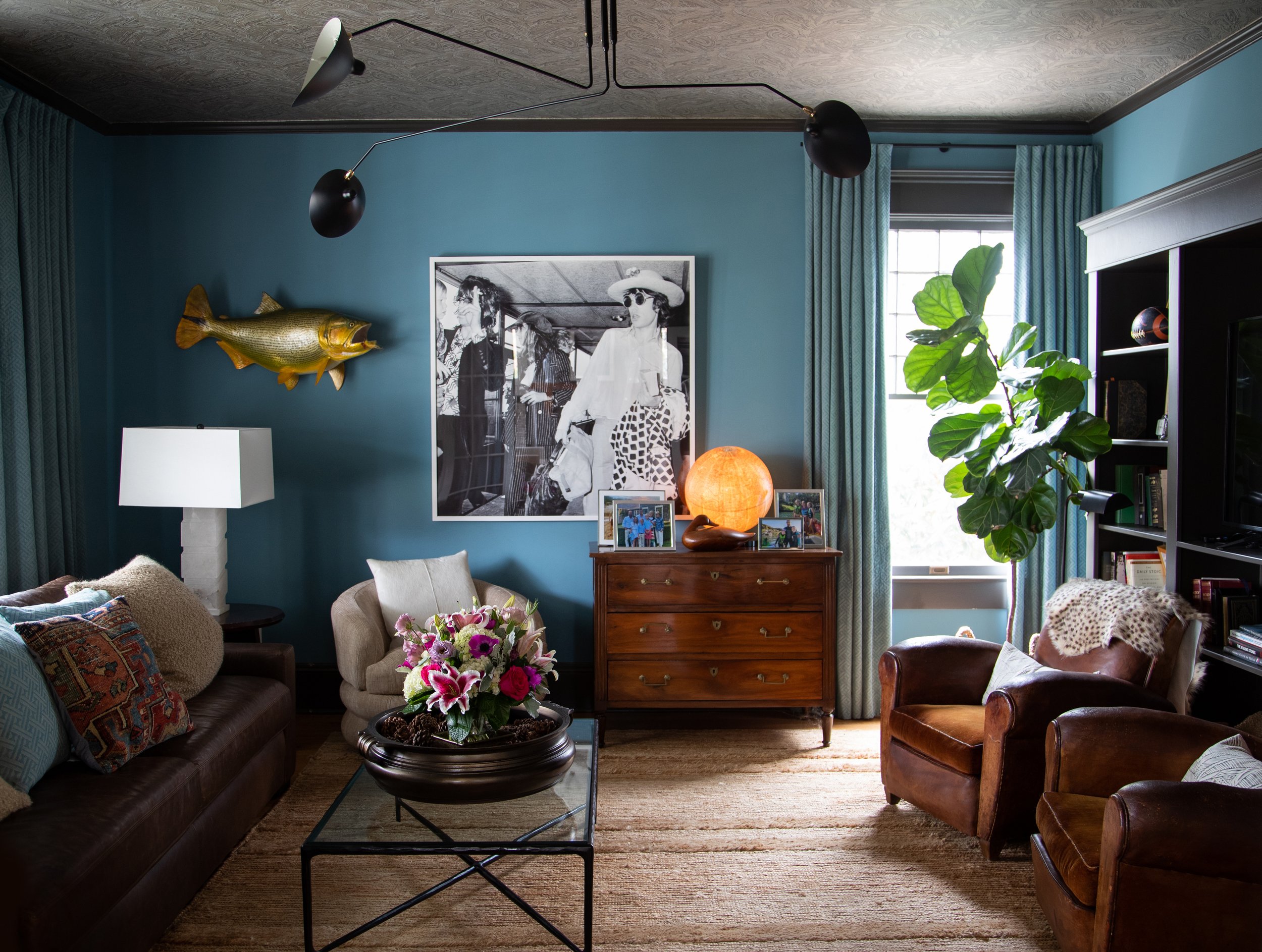 Living room with blue walls, brown leather furniture, a glass coffee table with a floral arrangement, wall-mounted fish decor, a large black-and-white photo, a wooden dresser, a fig tree, and a modern black ceiling light.