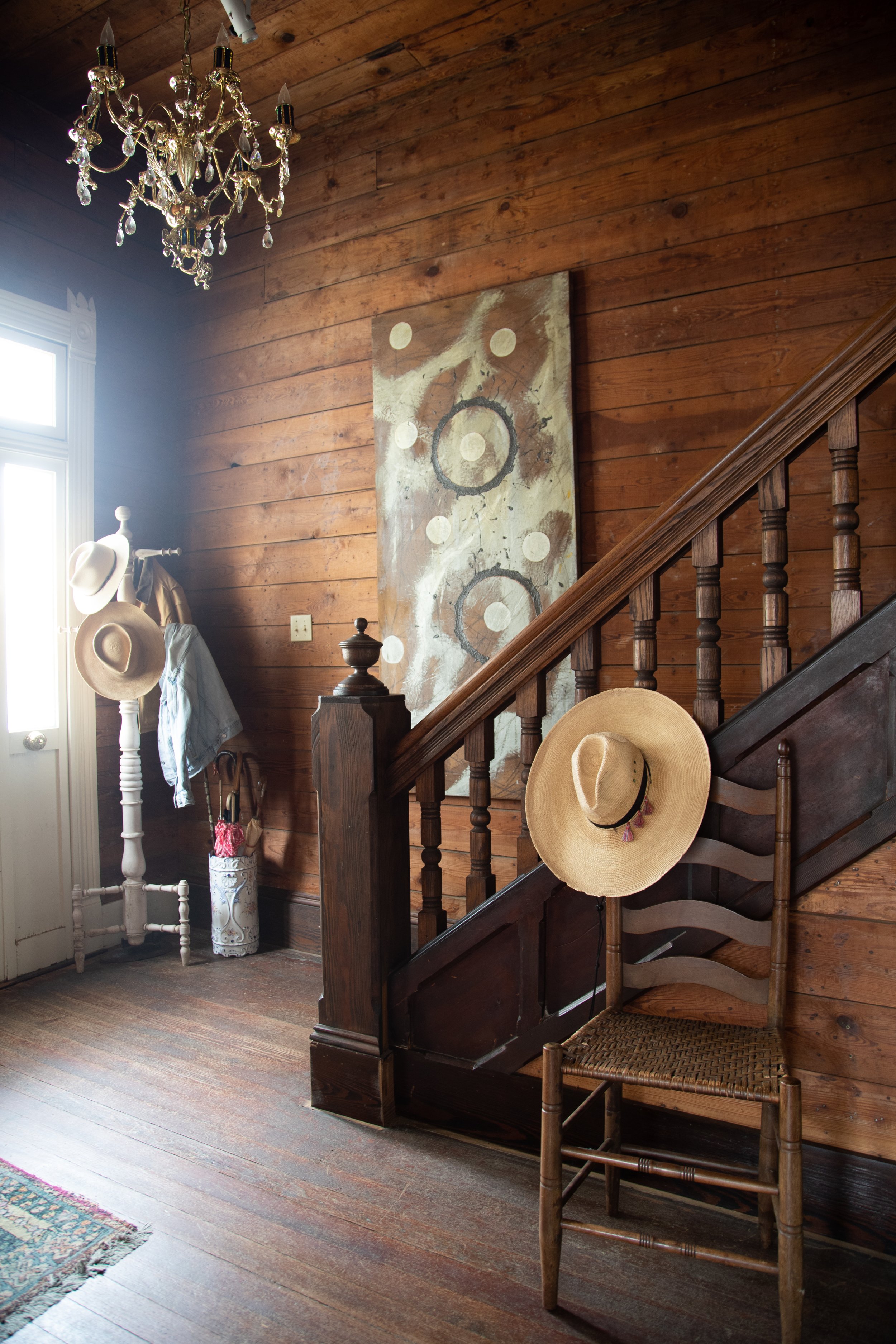 Rustic wooden interior with a staircase, ladder-back chair with a straw hat, abstract wall art, hat rack with jackets and hats, and a chandelier.