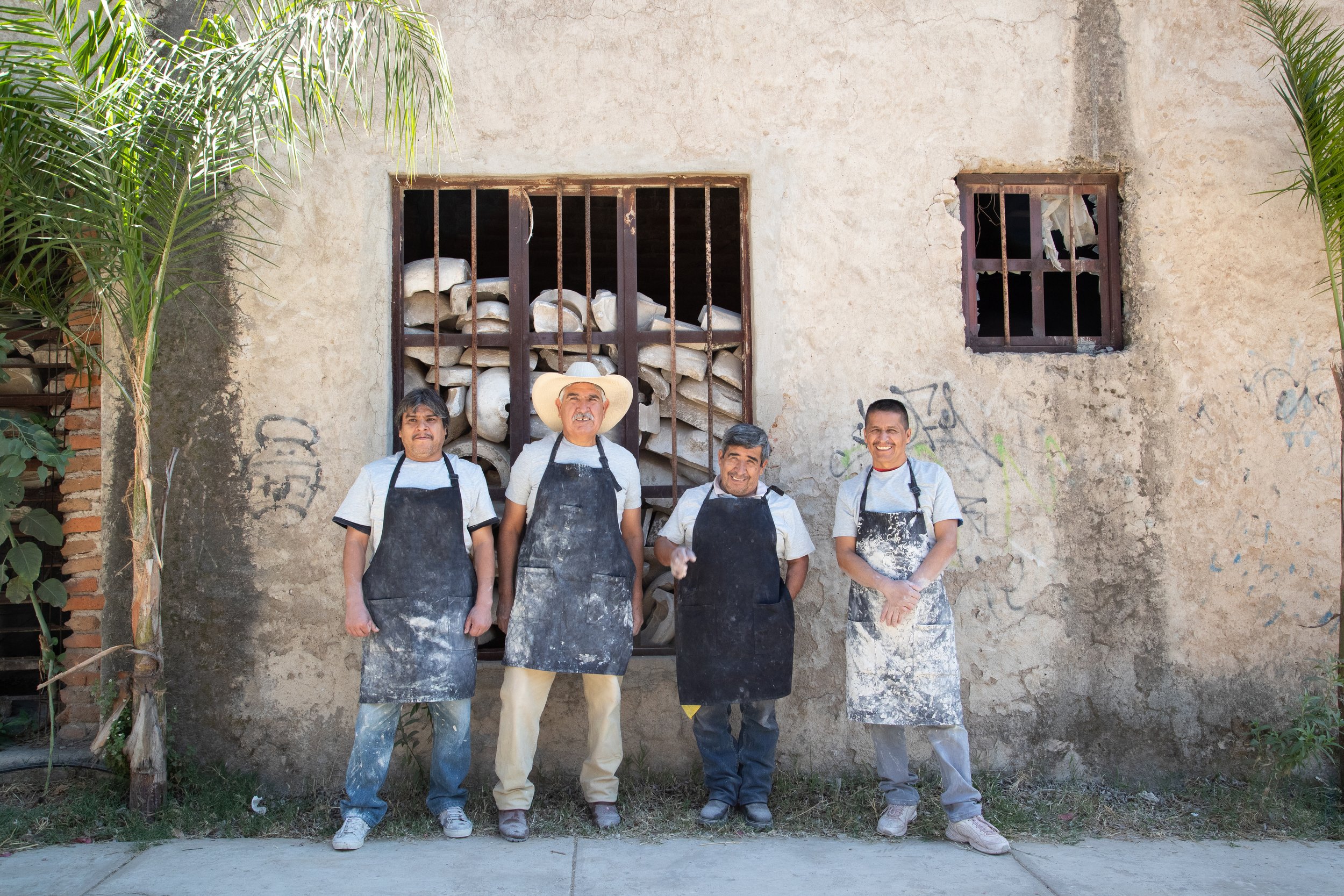 Four men in aprons standing outside a rustic building with a window filled with clay molds.