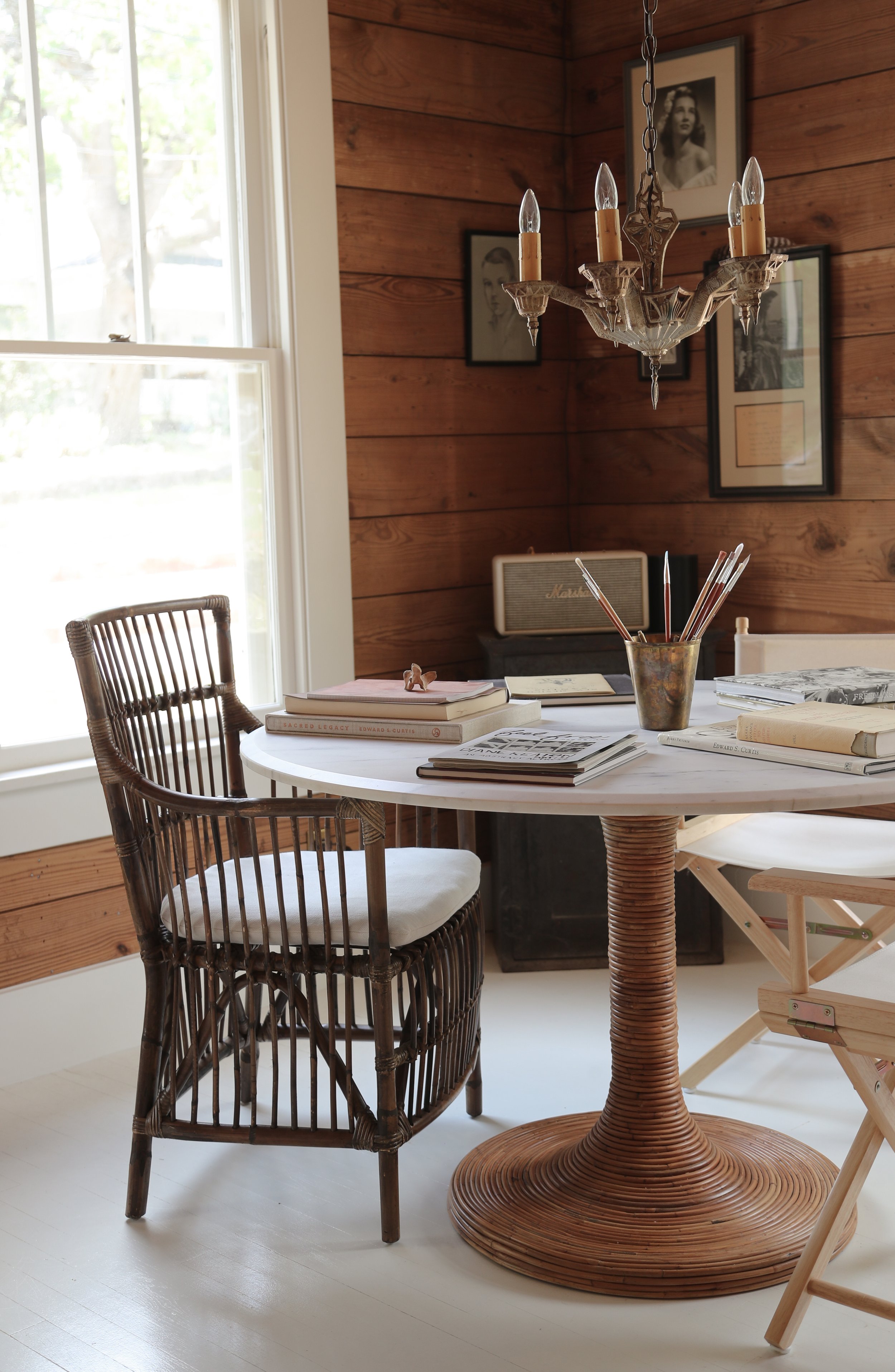 Room with wooden walls, a round table with books and pencils, wicker chairs, and a vintage chandelier.