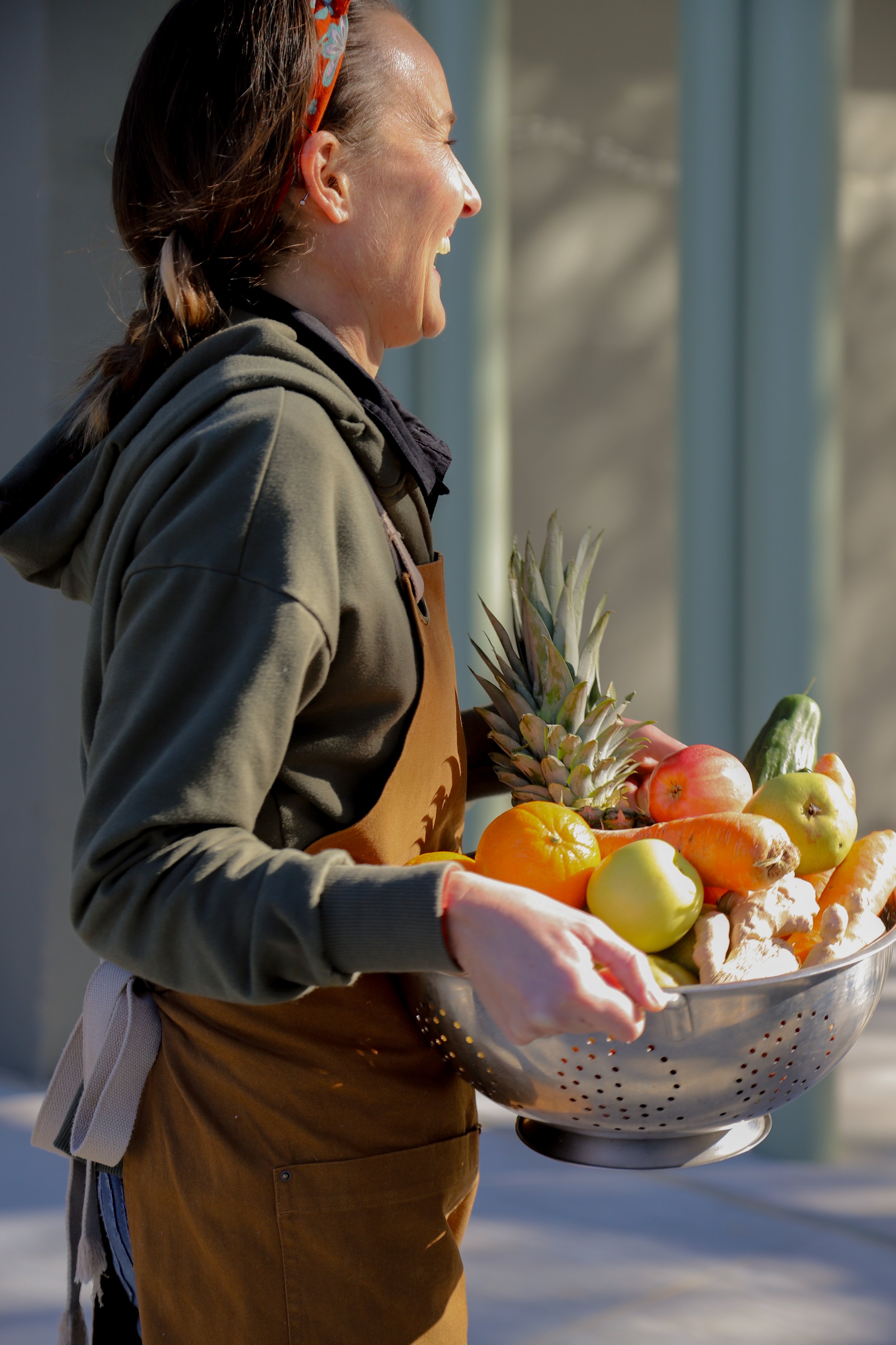 A woman holding a large bowl of fresh fruits and vegetables, including pineapple, apples, carrots, ginger, and tomatoes, standing outdoors in bright sunlight.