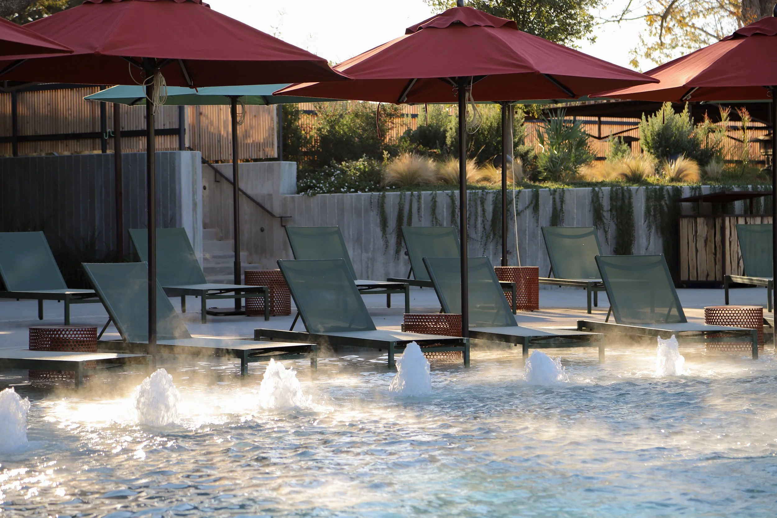 Empty poolside area with lounge chairs, large red umbrellas, and water fountains in the pool, surrounded by greenery and a wooden fence.