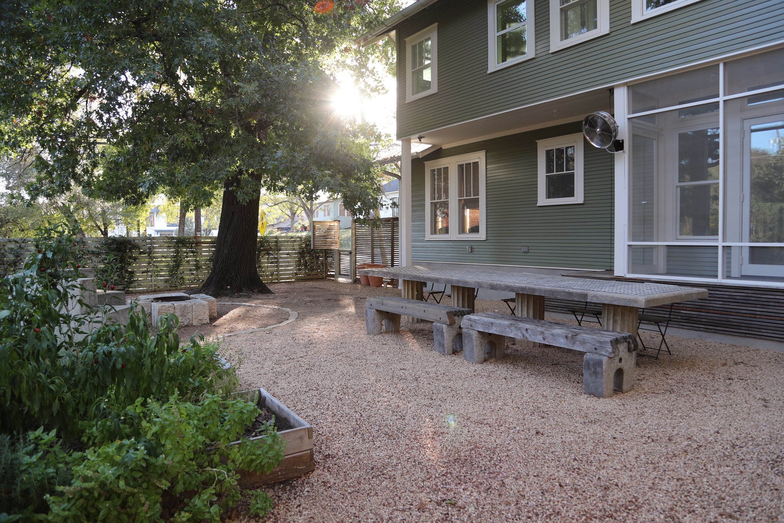 Backyard with wooden table and benches, large tree, garden bed, gravel ground, and green house exterior.