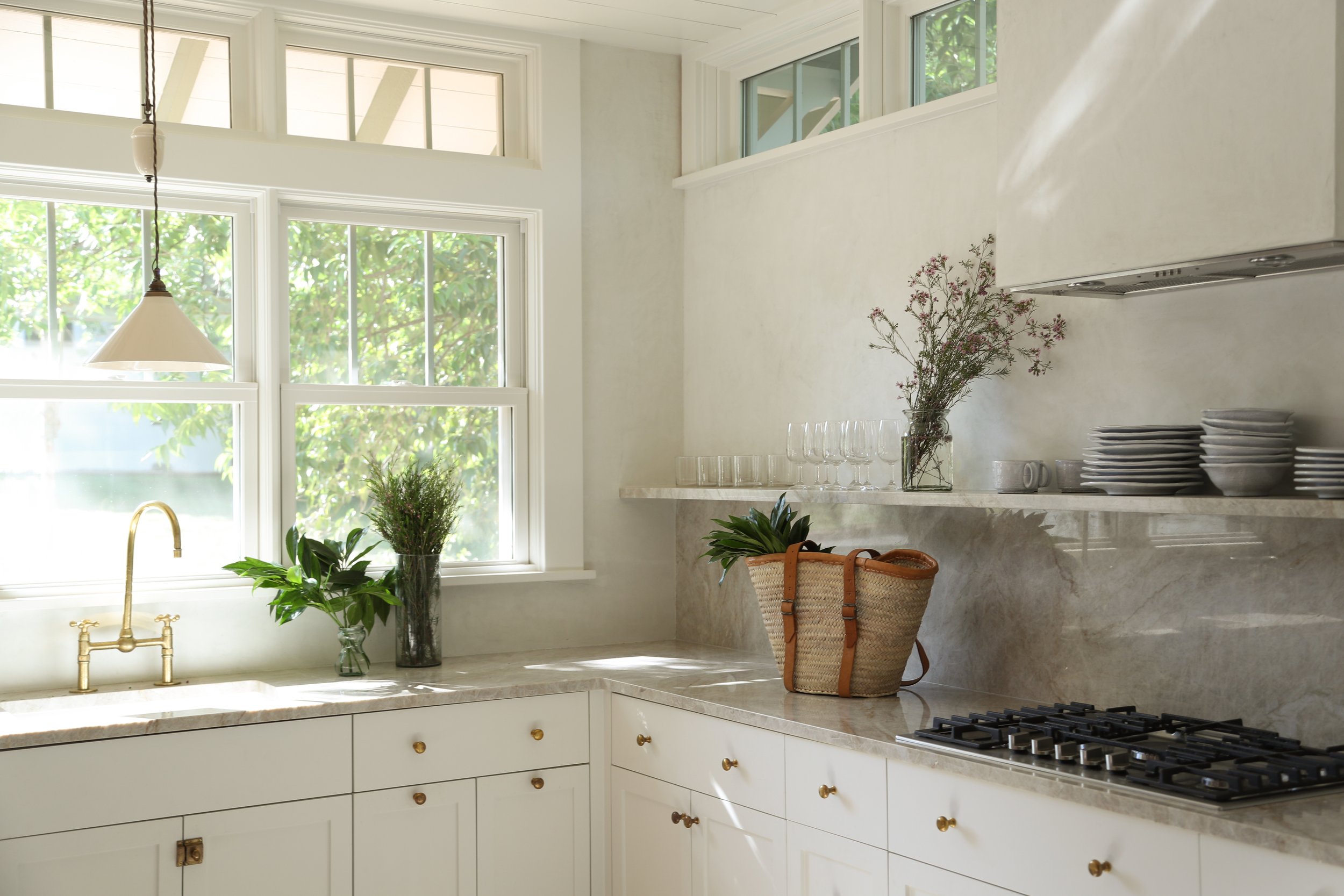 Bright kitchen with marble countertops, potted plants, wicker basket, gas stove, and open shelving with glasses and plates.
