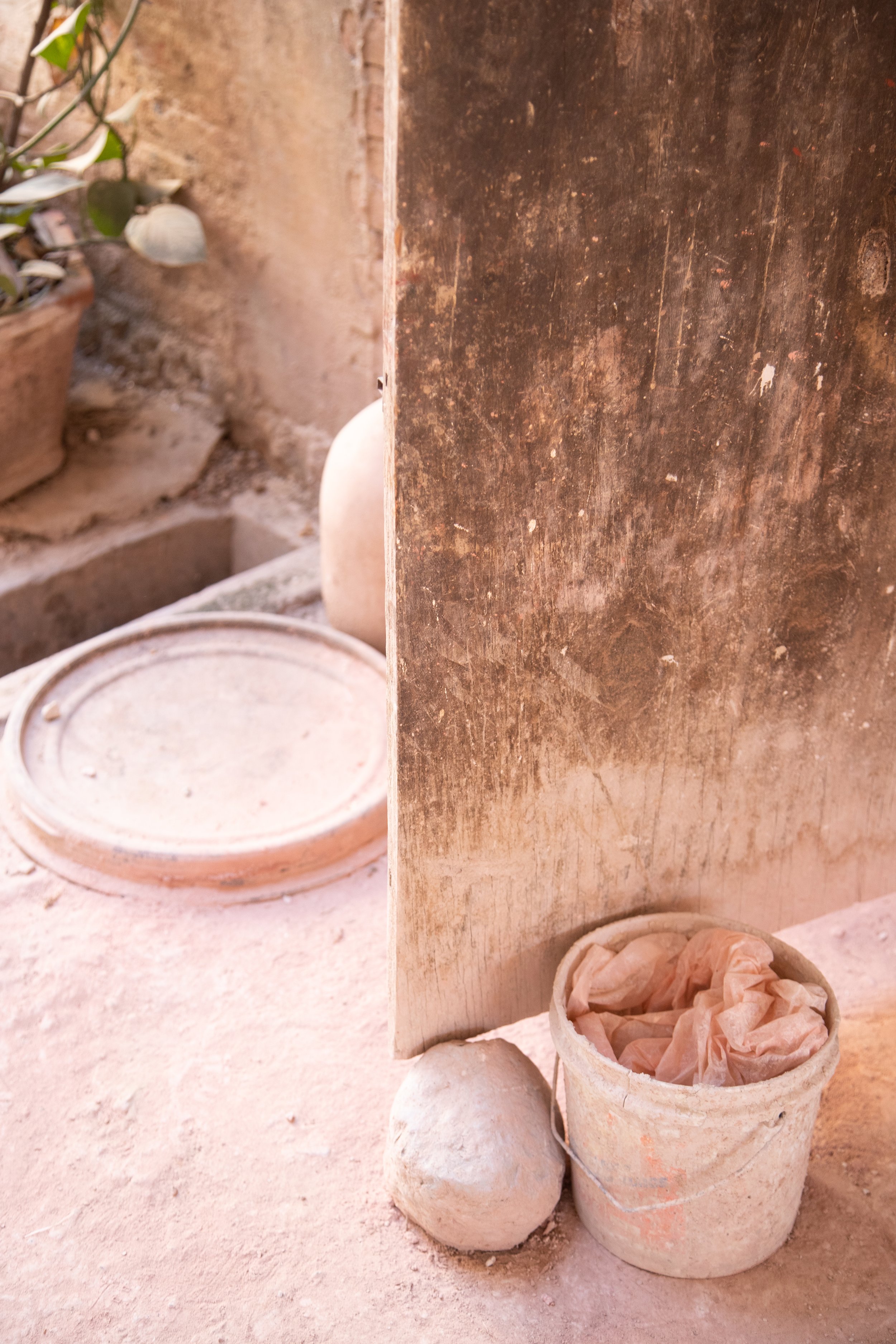 Dusty old wooden door slightly ajar with a bucket filled with crumpled paper nearby, resting on a dusty concrete floor. A round stone is visible near the bucket, with a faintly visible plant in a pot in the background.