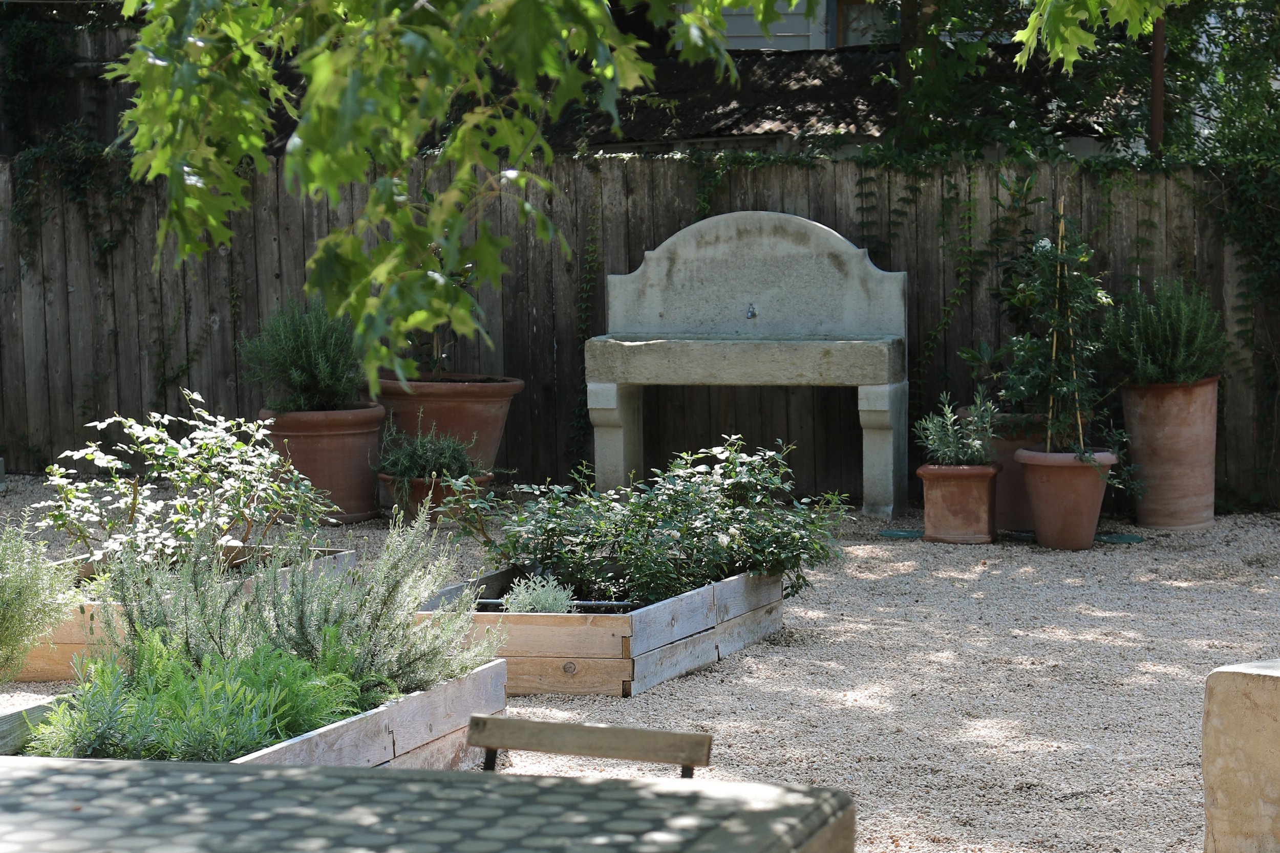 Outdoor garden with wooden planters filled with various plants and herbs, large terracotta pots, a stone garden sink against a wooden fence, and trees in the background.