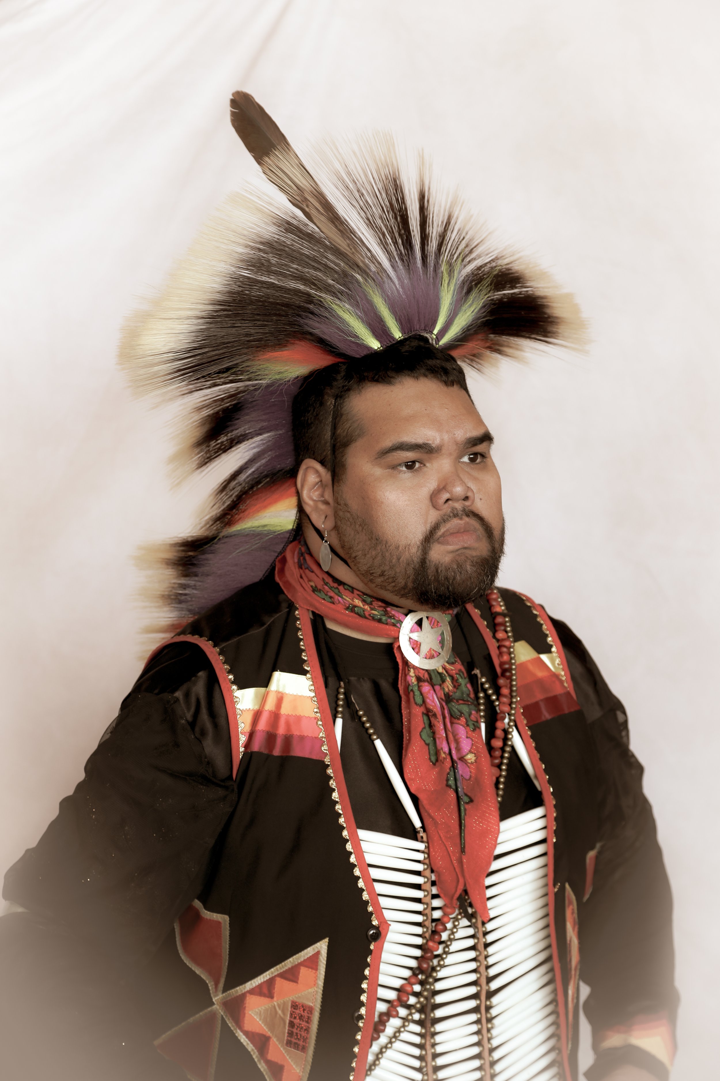 Person wearing traditional Native American regalia with a feather headdress, red scarf, and decorative jewelry, posing against a neutral background.