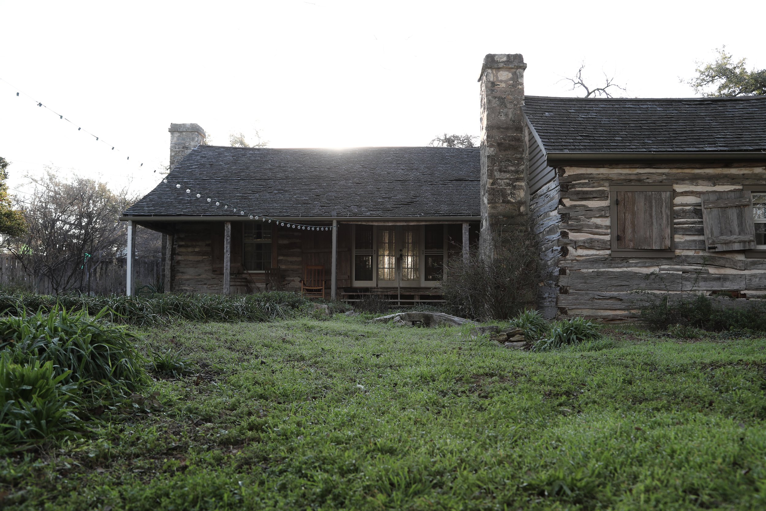 Historic rustic log cabin with stone chimney, surrounded by green grass, trees, and string lights on the porch in early morning light.