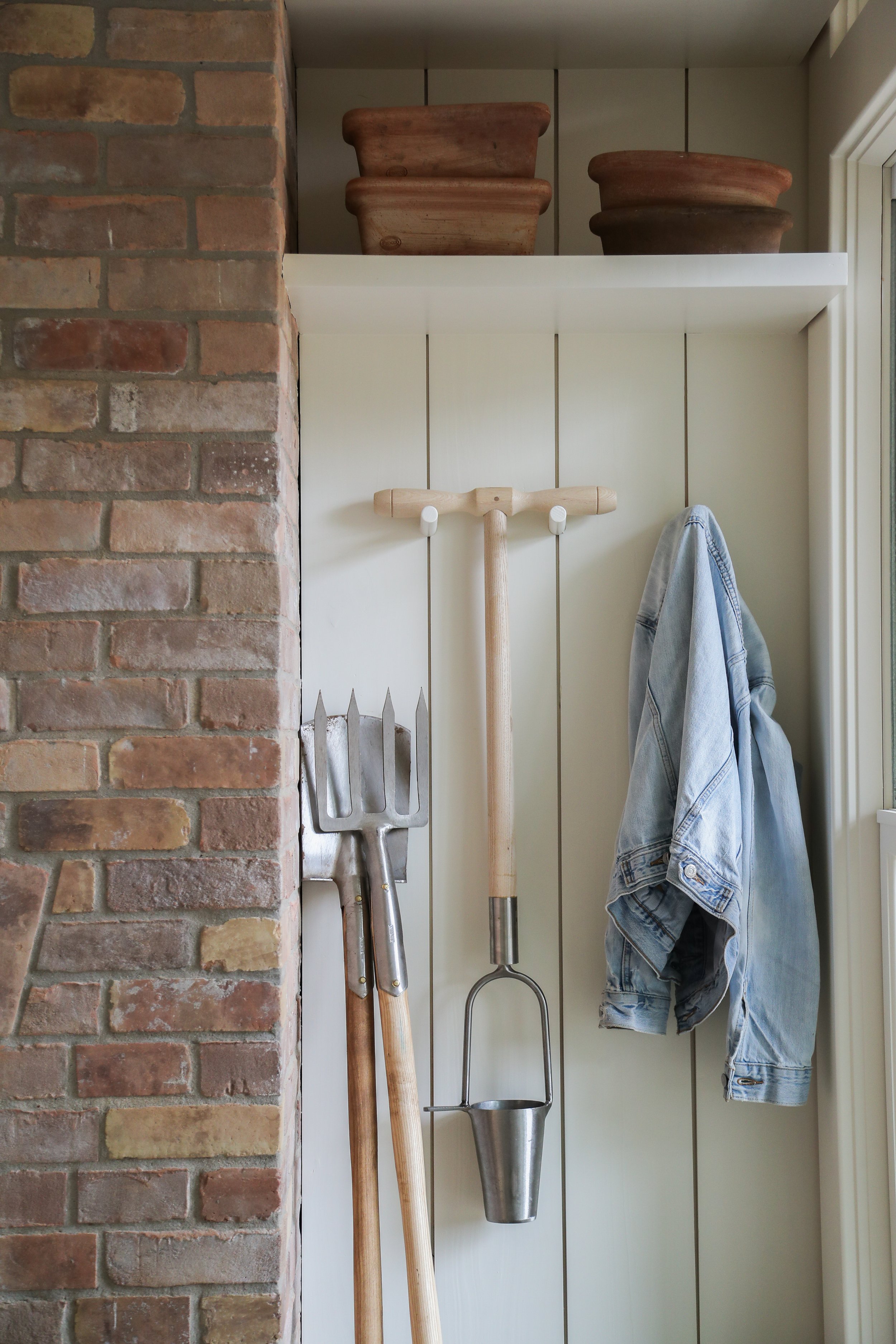Rustic corner with brick wall, gardening tools, denim jacket, and terracotta pots on a shelf.