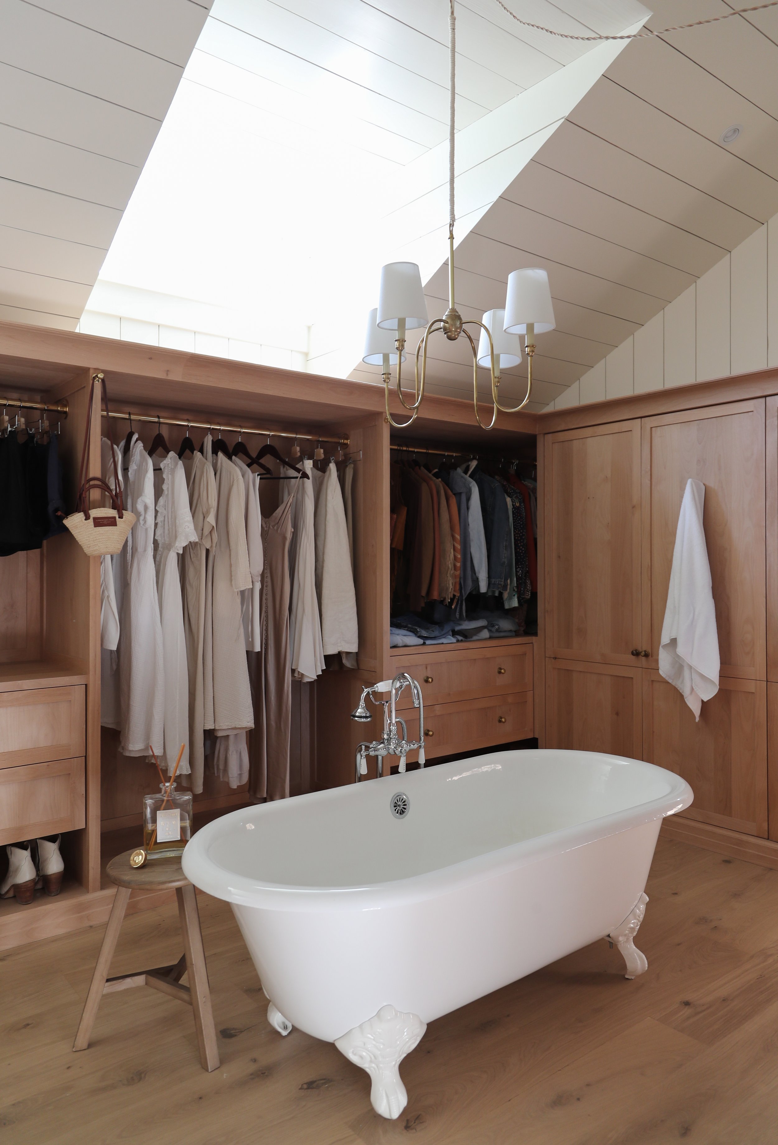 A cozy bathroom and closet space with a freestanding white clawfoot bathtub in front of an open wardrobe. The wardrobe is filled with neatly hanging clothes in light and neutral colors. A small side table with a candle and diffuser is beside the bath