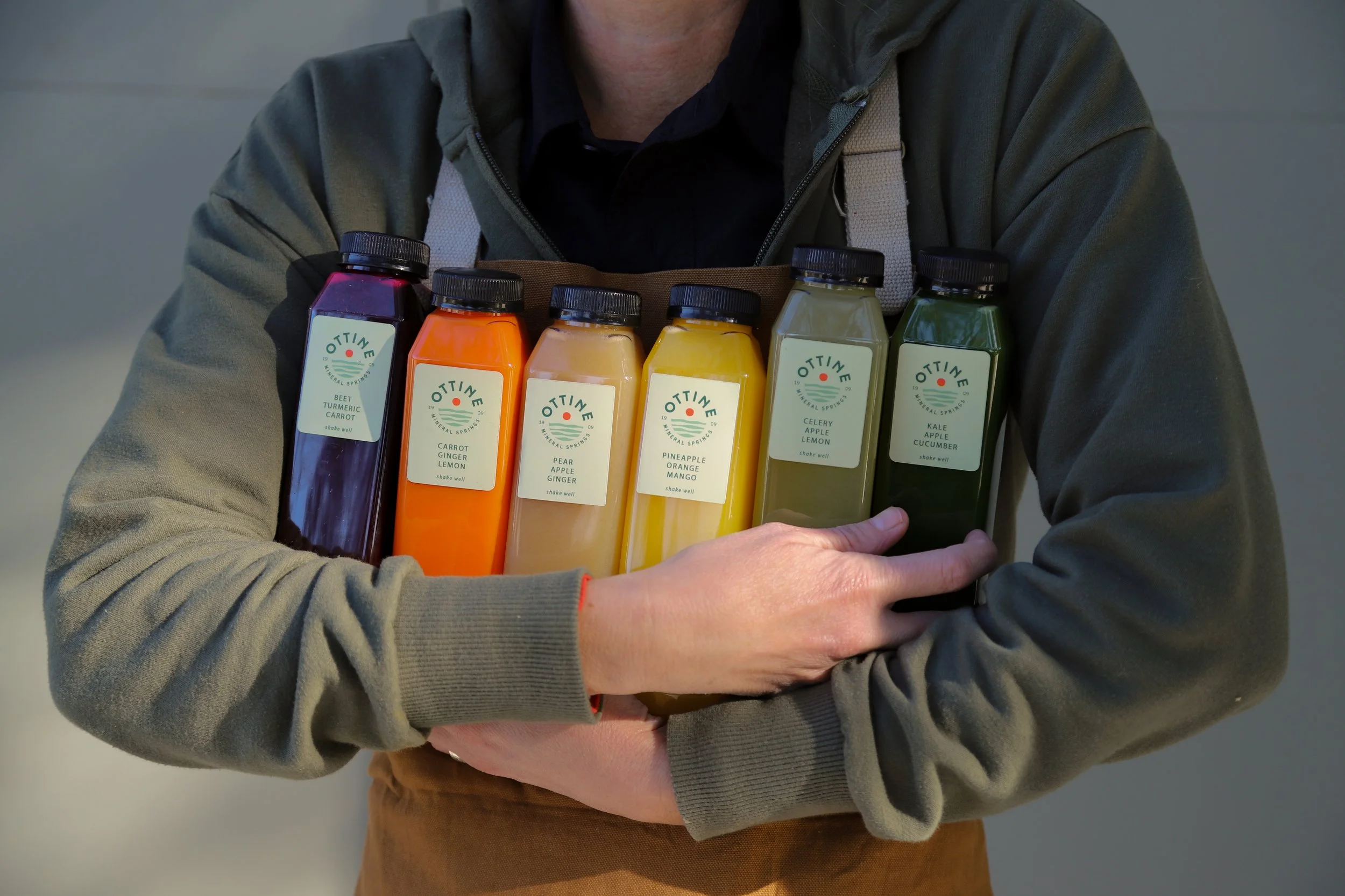 Person holding bottles of colorful vegetable and fruit juice, labeled with flavors such as turmeric carrot, carrot ginger lemon, pear apple ginger, pineapple orange mango, celery apple lemon, and kale apple cucumber.