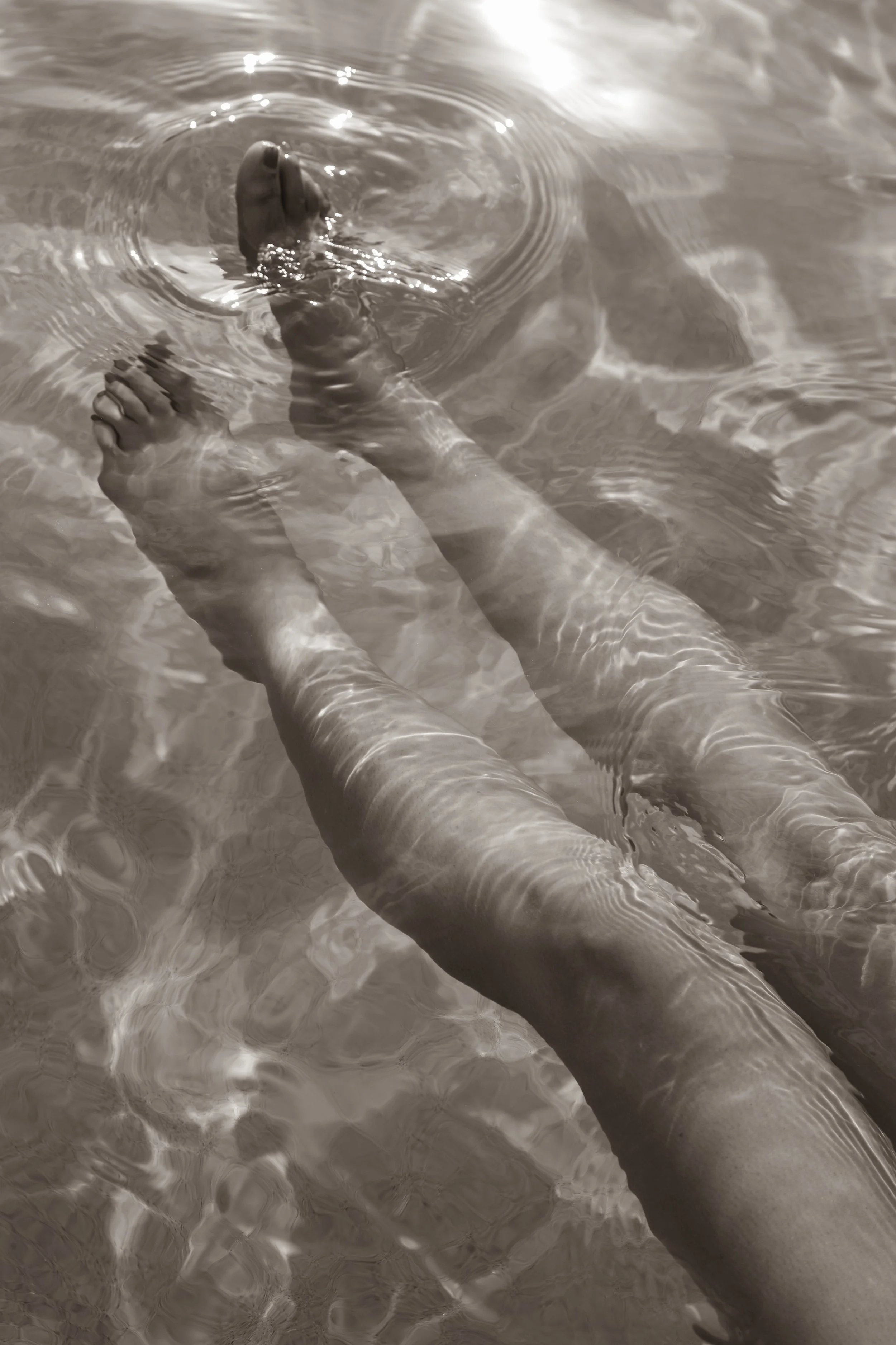 A person submerged in water up to their knees, showing their legs and feet, with ripples on the water's surface.