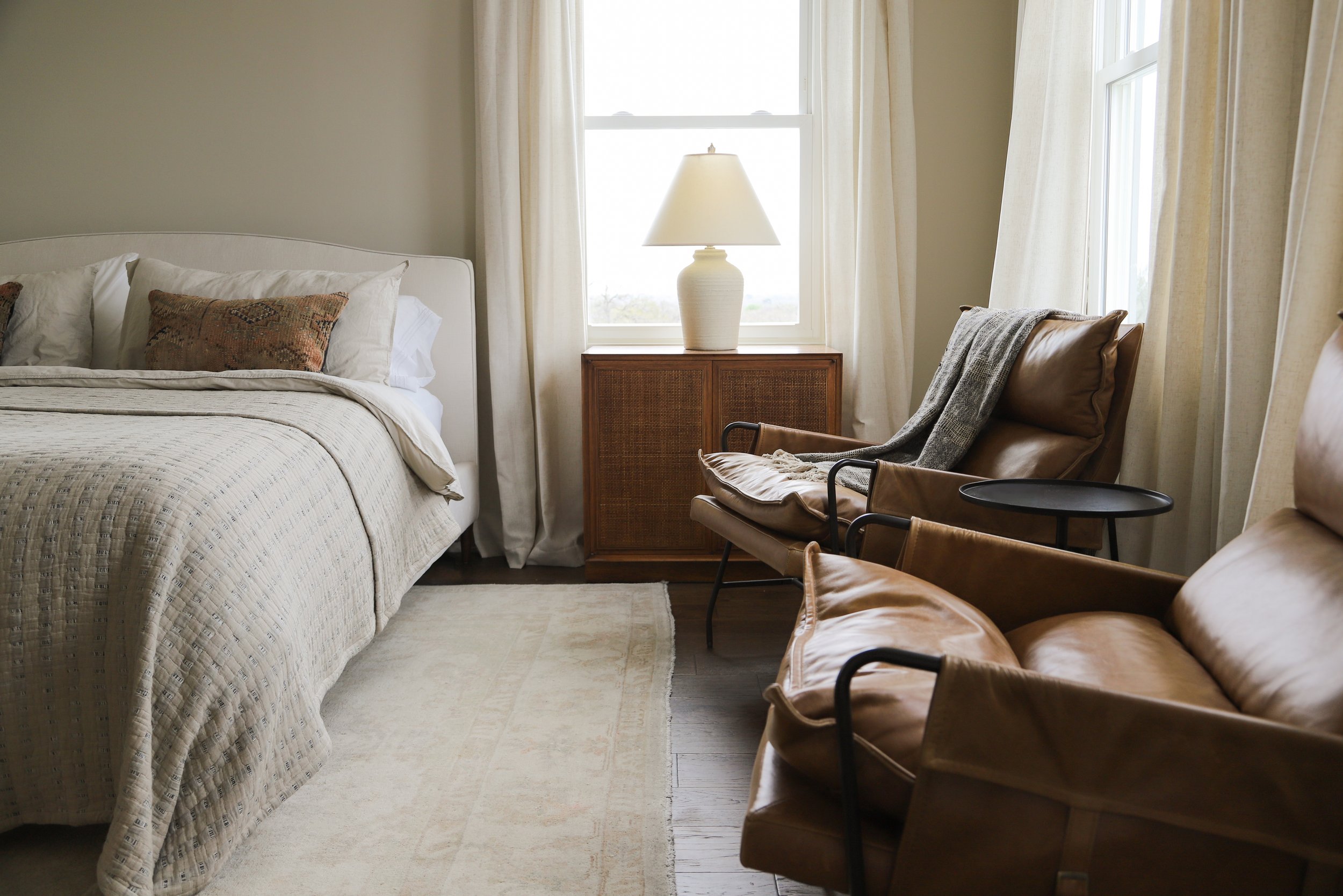 A bedroom with a white bed, beige quilt, pillows, a wooden cabinet with a lamp, two leather chairs, and a small black side table, illuminated by natural light from a window with cream curtains.