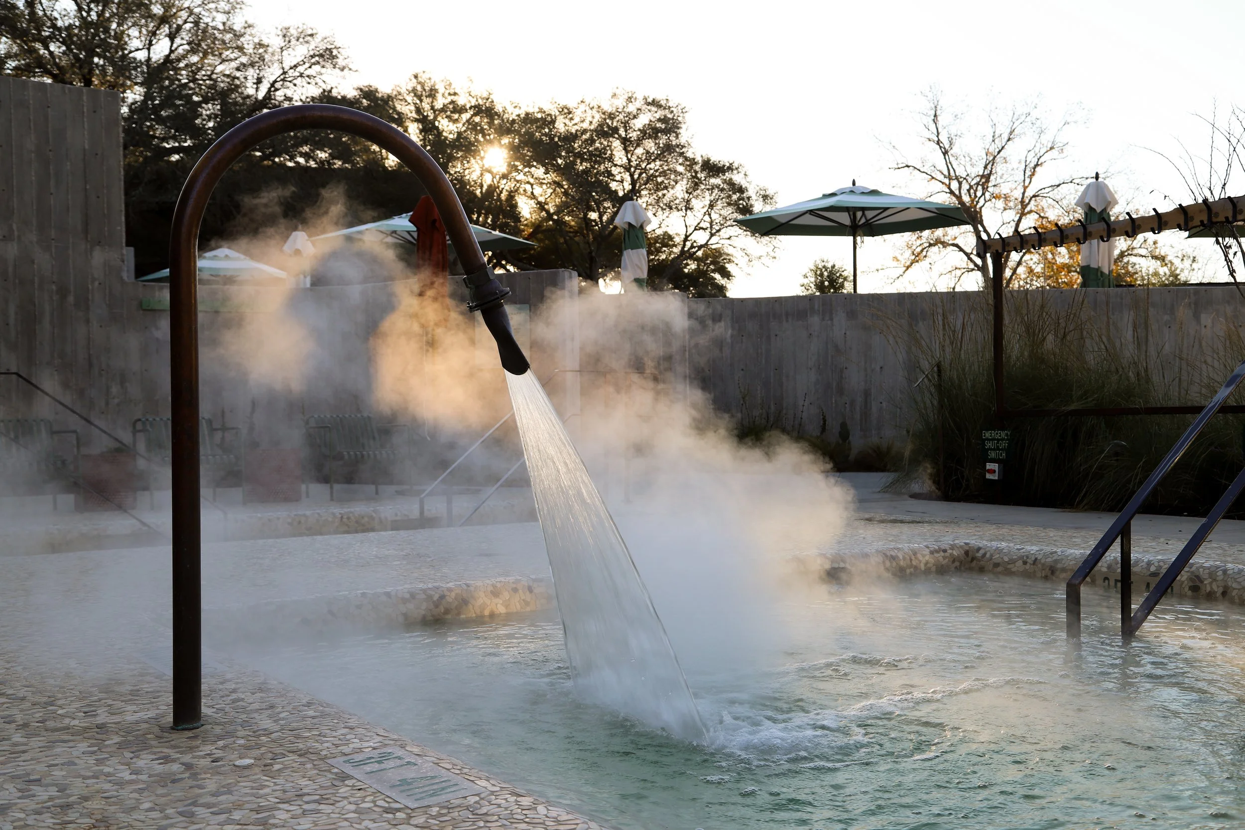 Outdoor hot spring or spa pool with steam rising, a shower head spraying water into the pool, pool steps on the right, and a wooden fence with umbrellas and trees in the background.