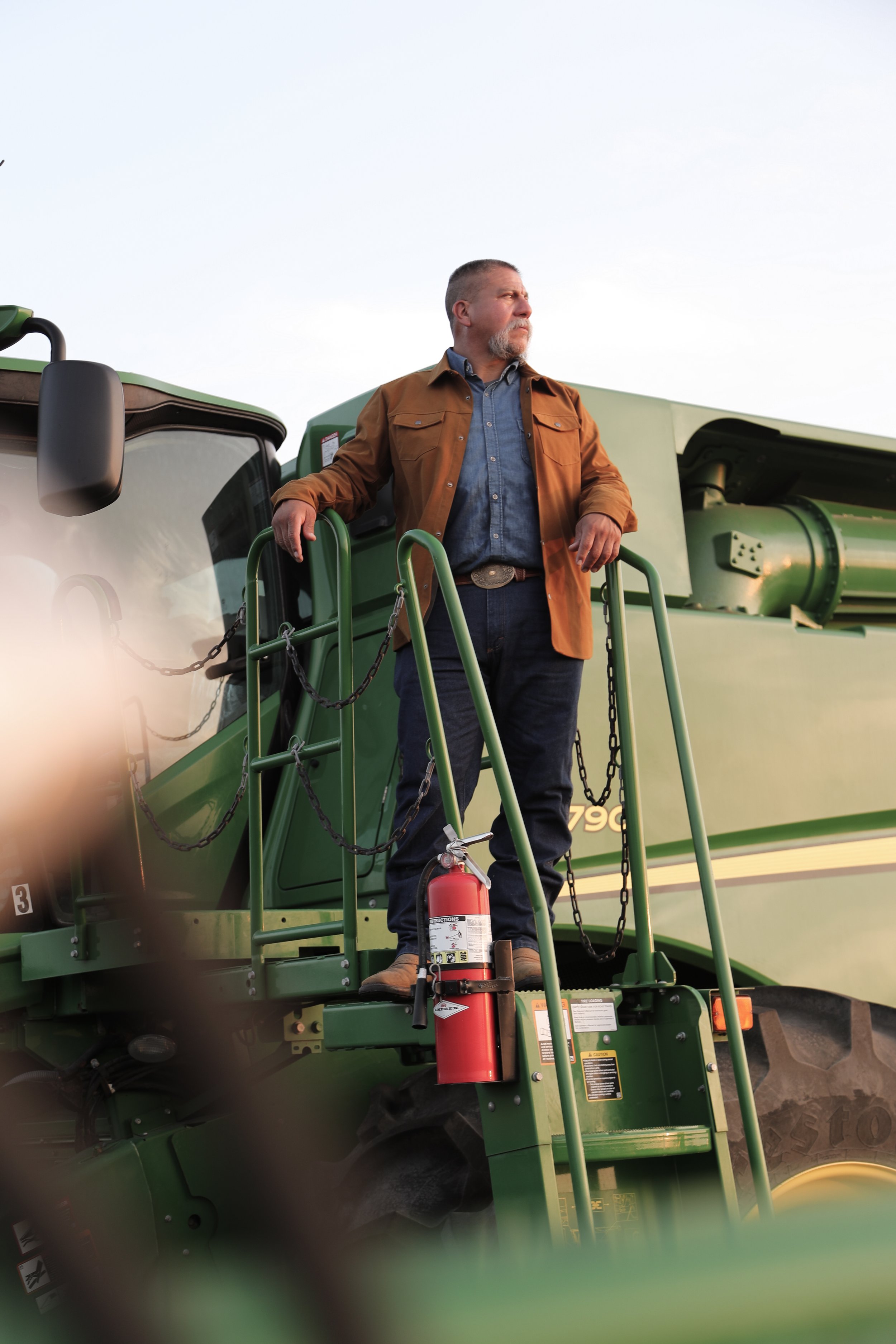 Man standing on a green agricultural machine, wearing a brown jacket and blue jeans, with a fire extinguisher attached to the machine.