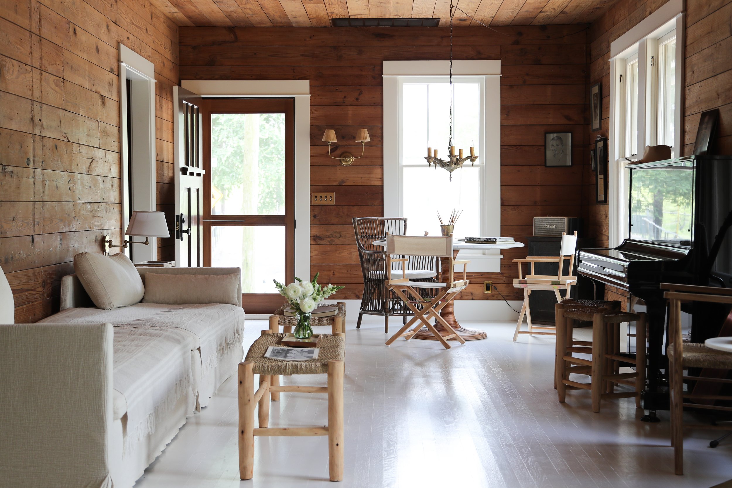 Rustic living room with wood paneling, beige sofa, wicker coffee table, dining area with director's chairs, and a piano with amplifier.
