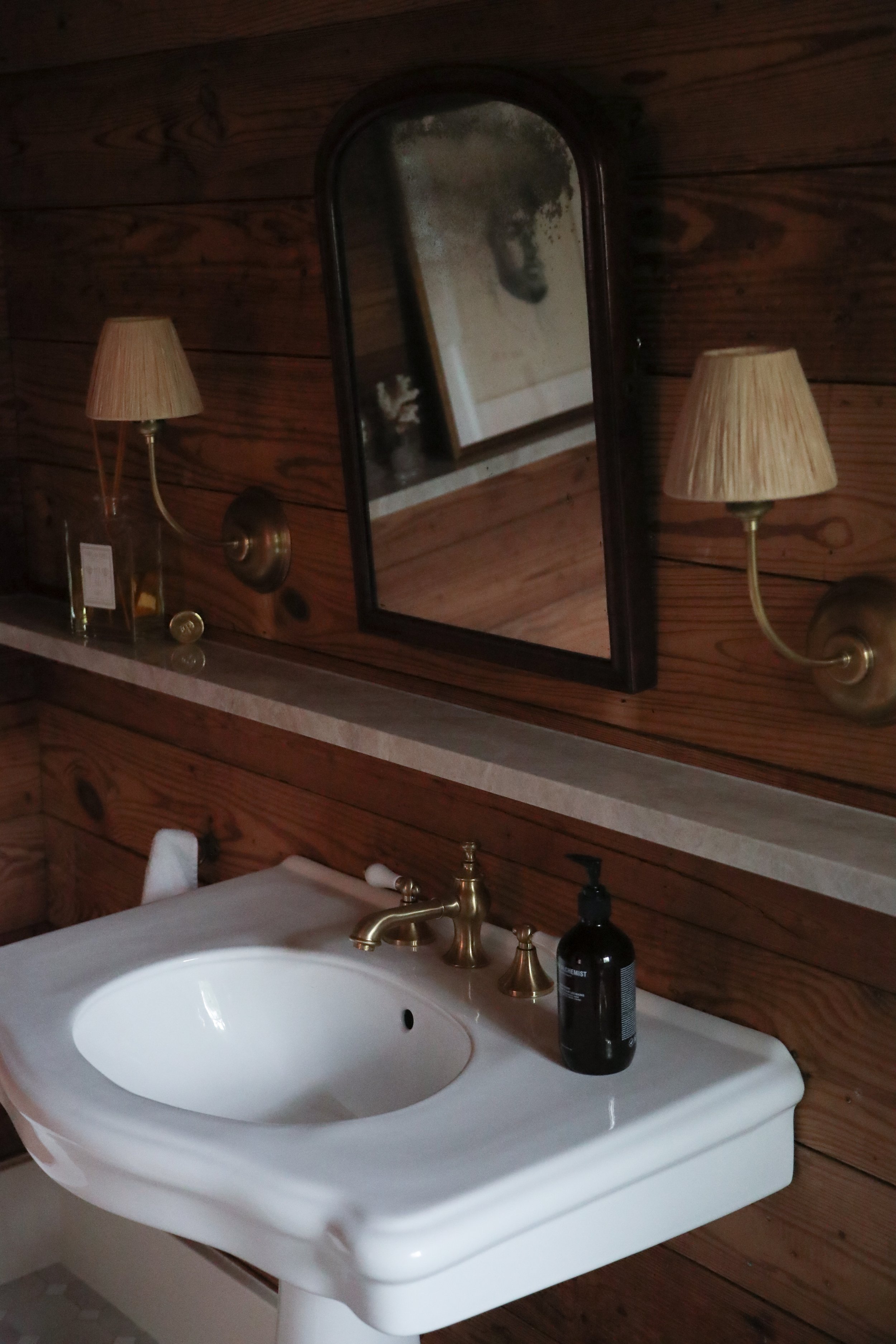 Rustic bathroom with wooden walls, a white pedestal sink, brass faucet, and a black soap dispenser. Antique mirror with reflection of a framed picture, and two wall-mounted lamps with fabric shades.