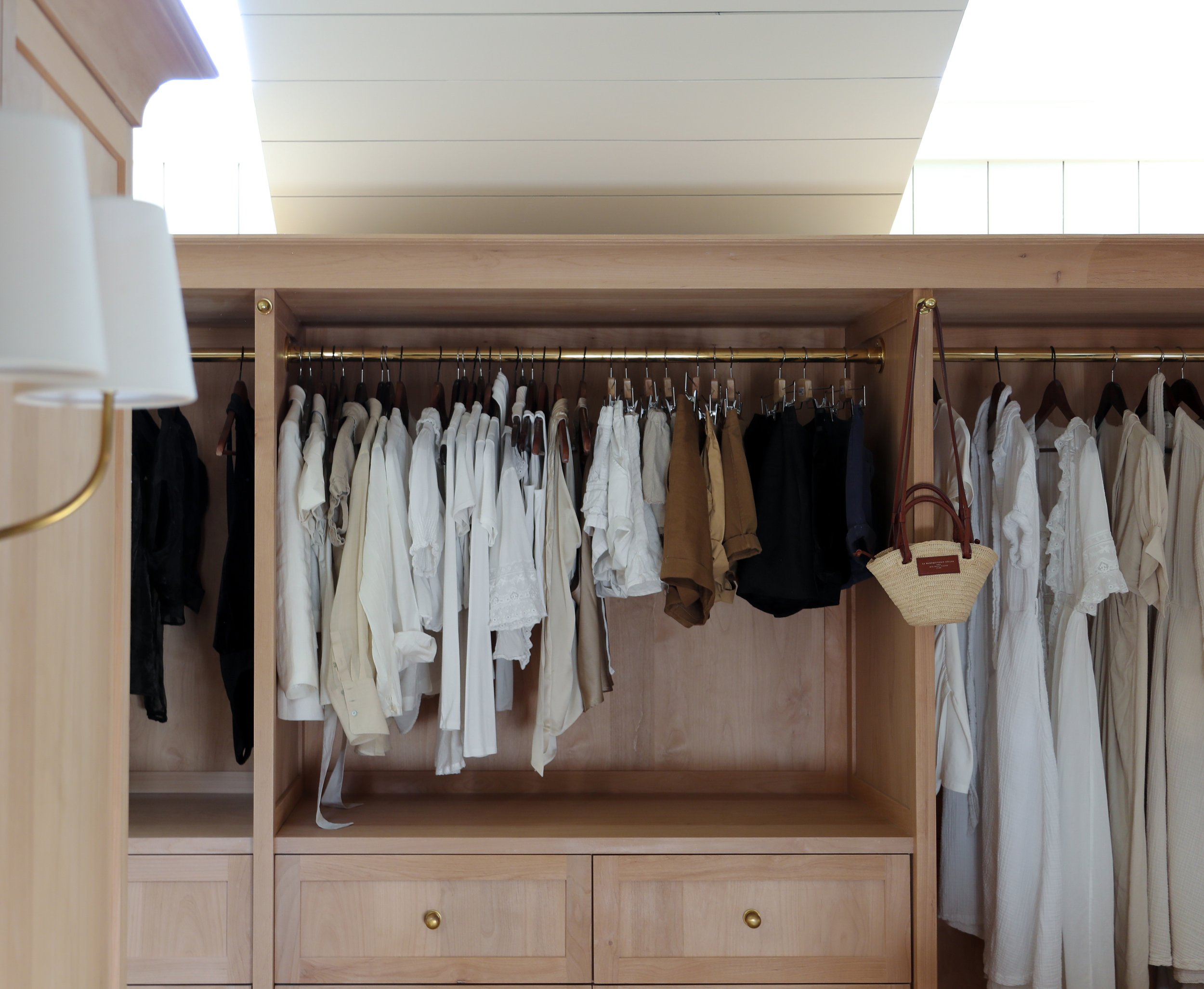 Open closet with neatly arranged clothes on hangers, mostly in white and neutral colors. A small straw handbag is hanging on a hook. The closet features wooden drawers below the clothing.