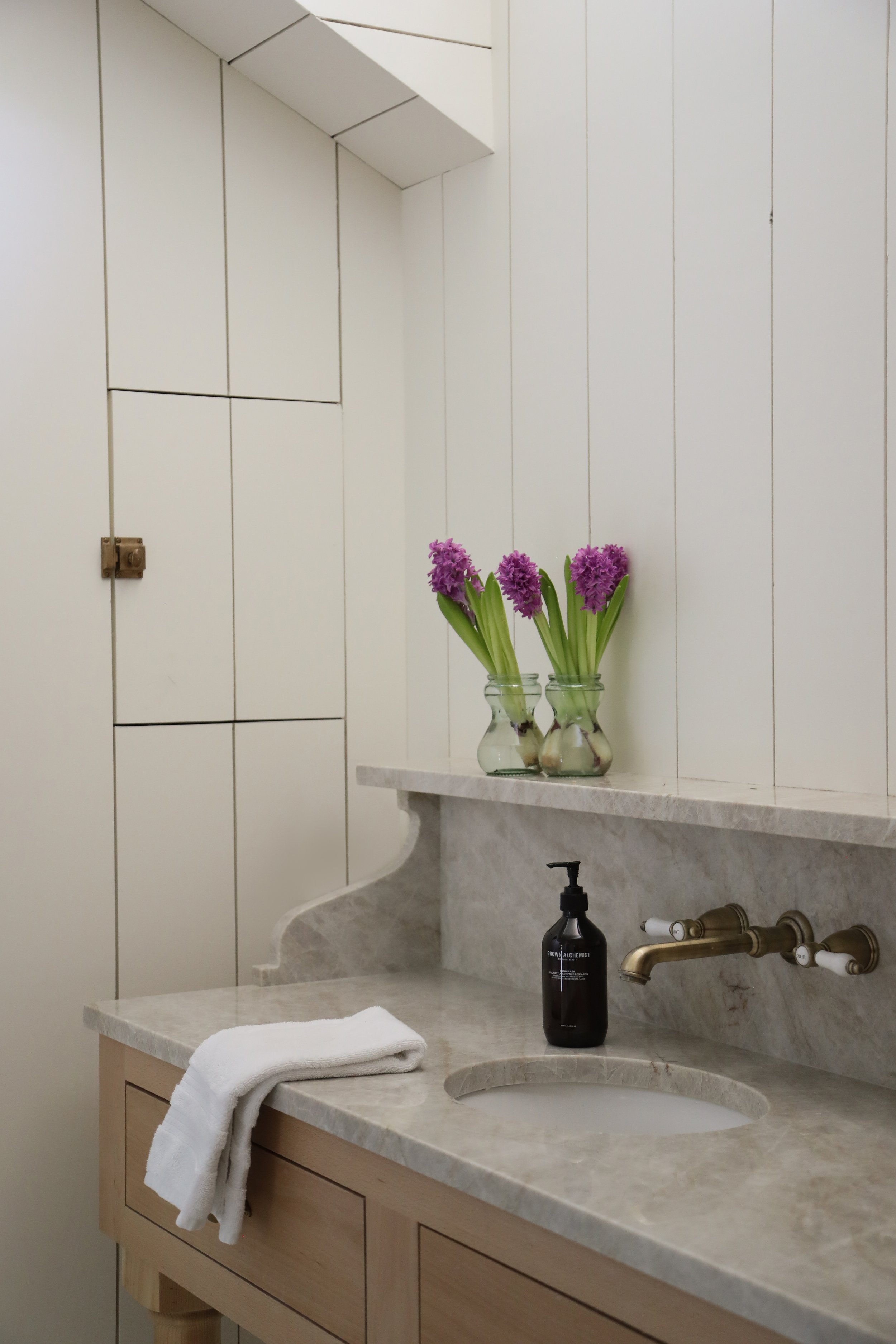 Bathroom vanity with marble countertop, brass faucet, folded white towel, black soap dispenser, and purple flowers in glass vases on a shelf.