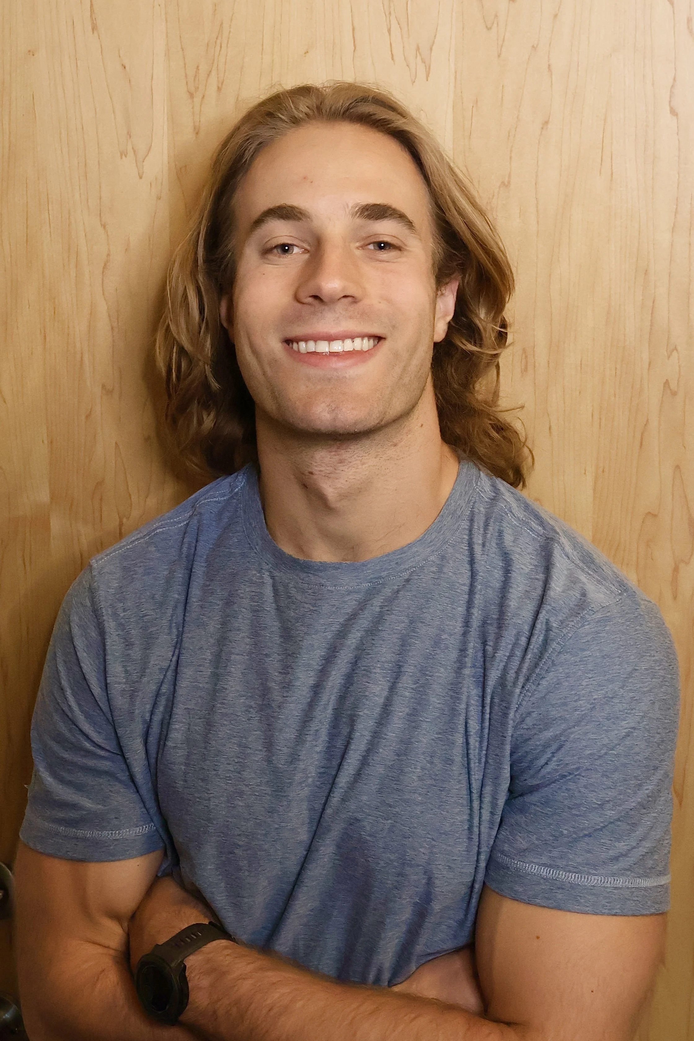 A smiling young man with long blond hair, wearing a blue t-shirt and a black watch, standing in front of a wooden wall.