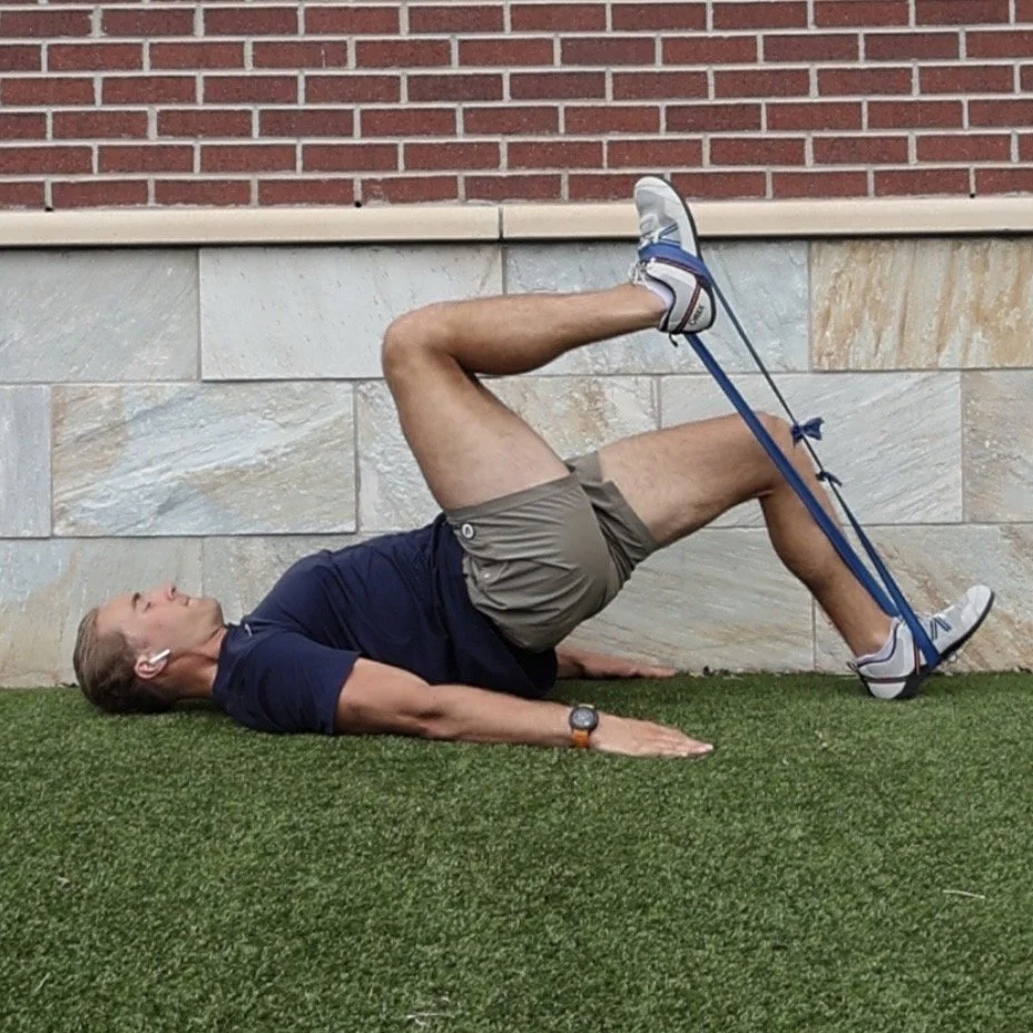 A man doing a leg exercise using a resistance band tied to a wall, lying on his back on a grassy surface.
