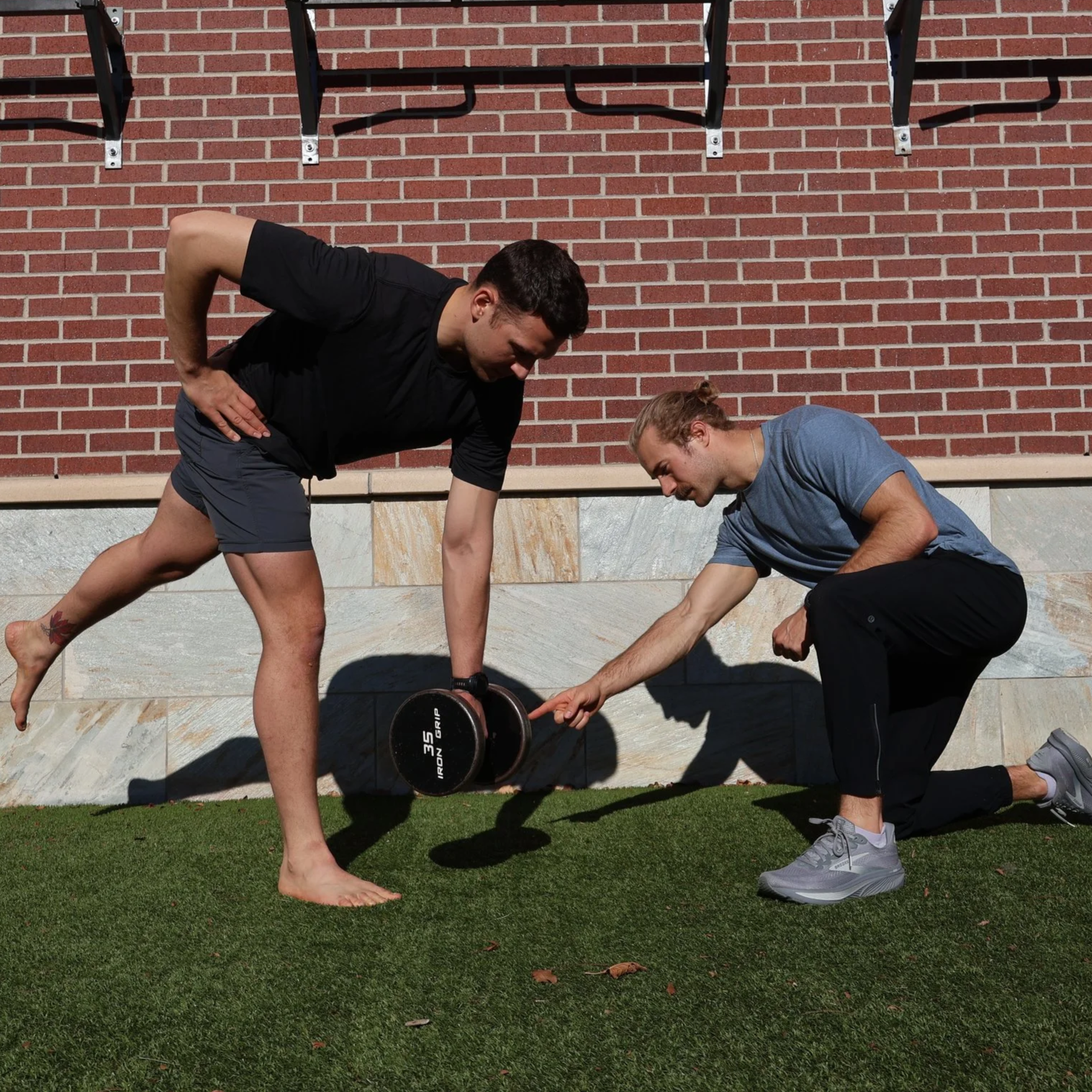 A man with a tattooed leg doing a balance exercise on one leg while a trainer supports him, outdoors in front of a brick wall, during a fitness training session.