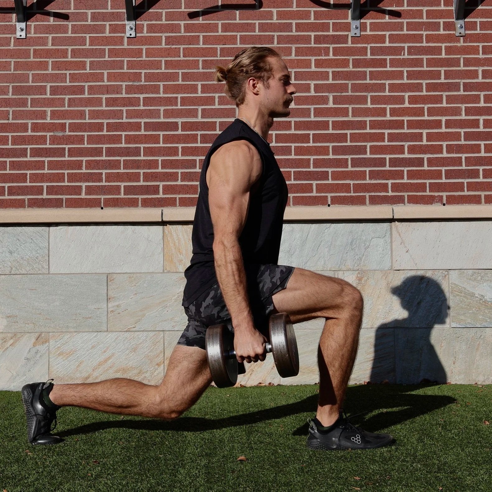 A man exercising outdoors with a dumbbell, lunging with his right leg forward on green grass against a brick wall.
